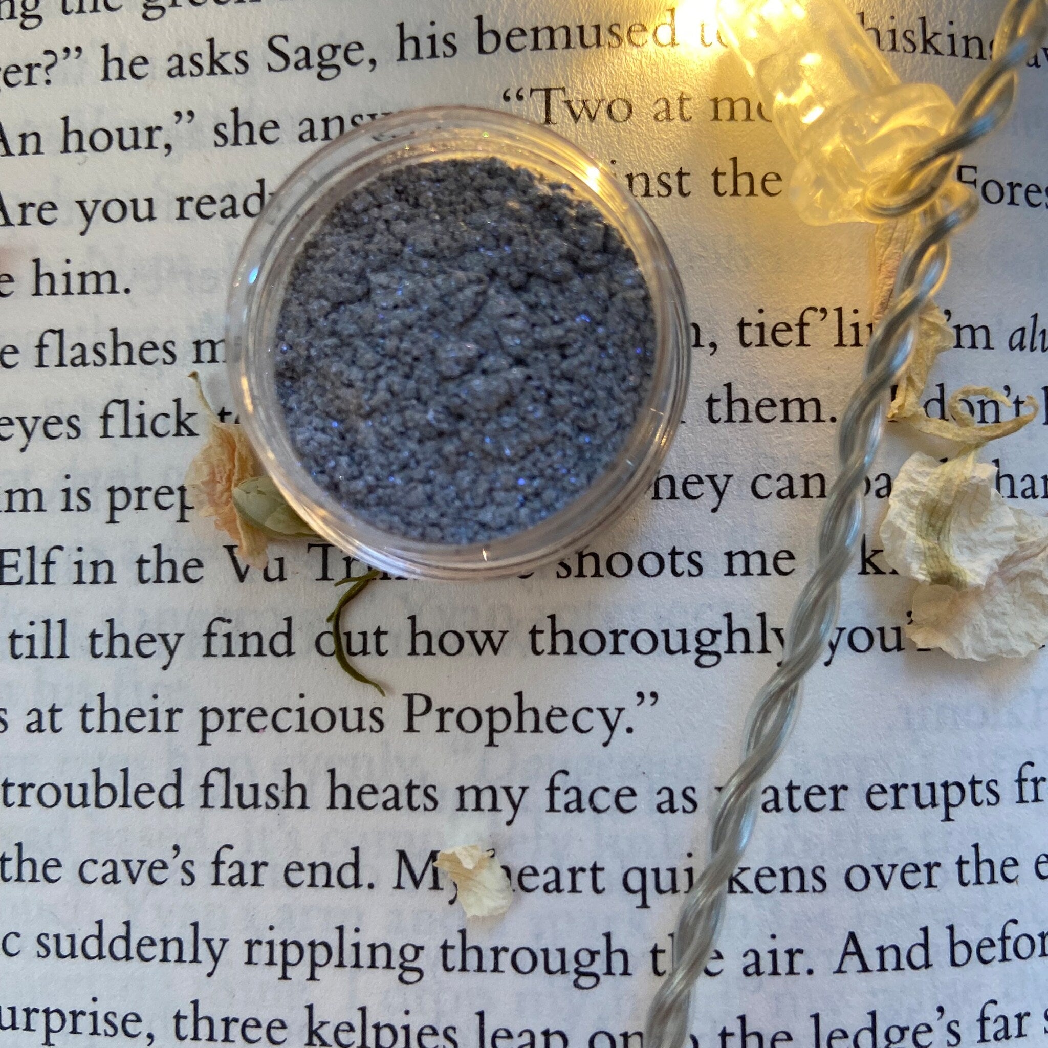Glitter eyeshadow in gray, sparkly, contained in clear plastic jar on book page