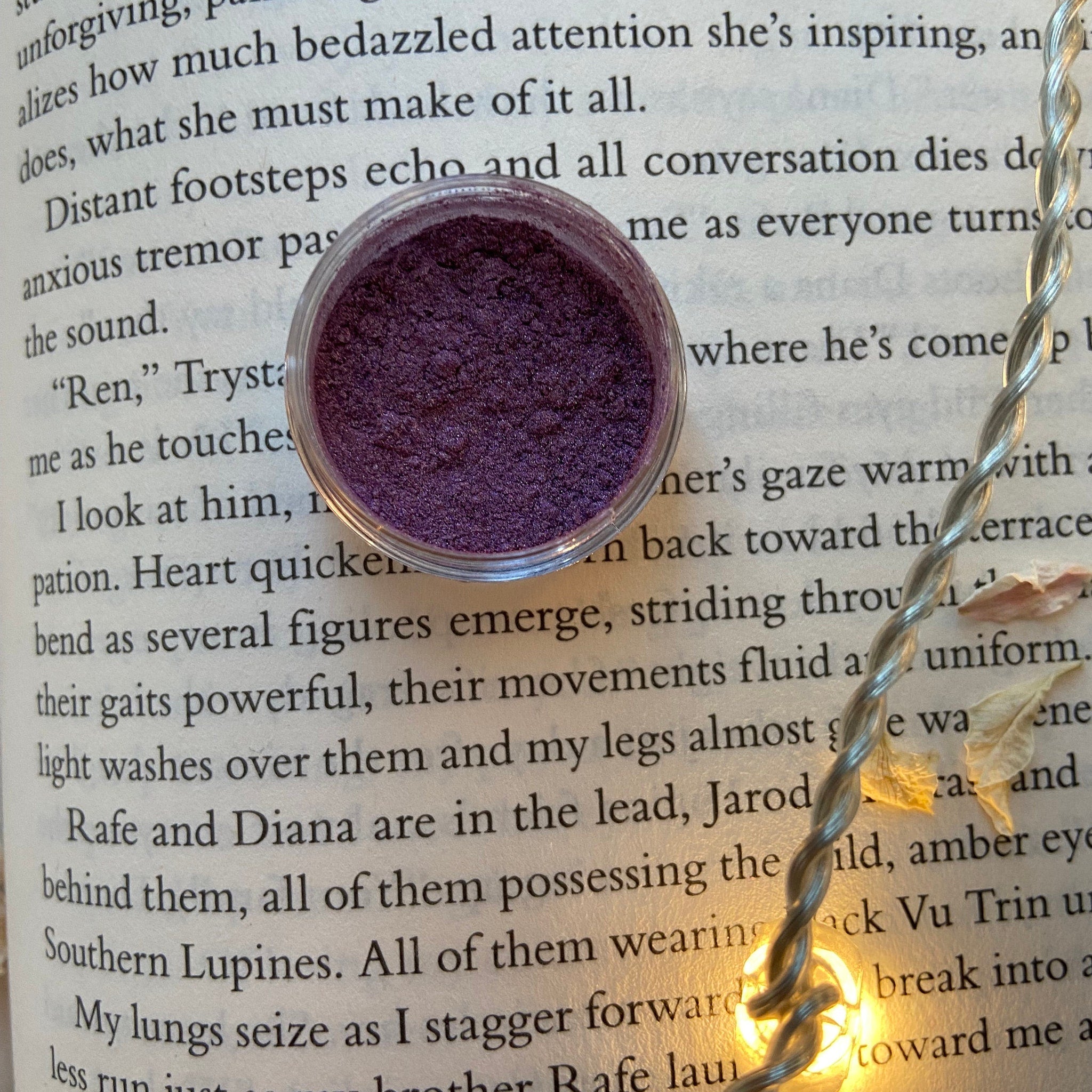 Eye shadow, dark purple powder in clear jar, on open book page, with fairy lights