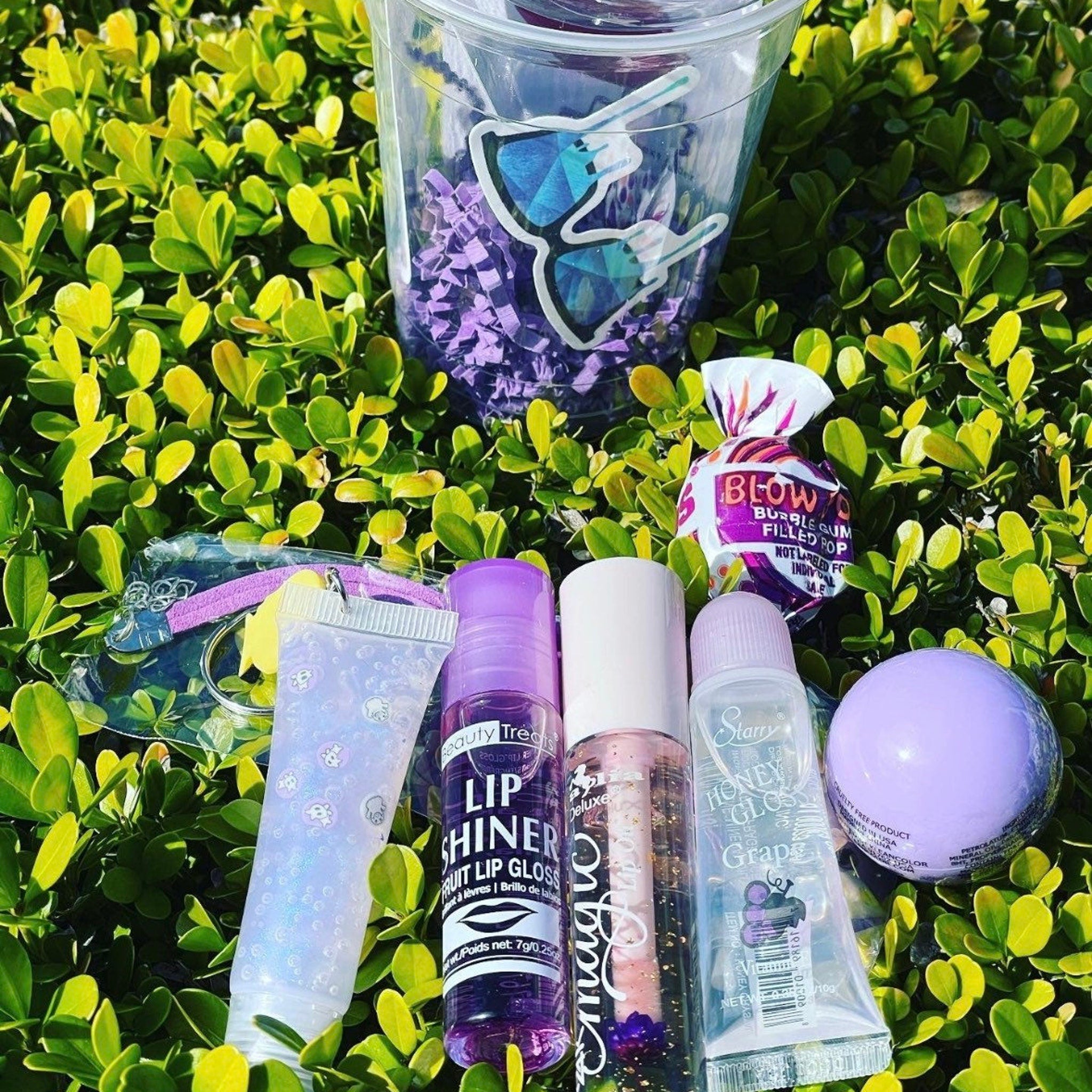 Lip gloss set with clear, purple, and pink tubes, plus a purple lip balm, on green leaves