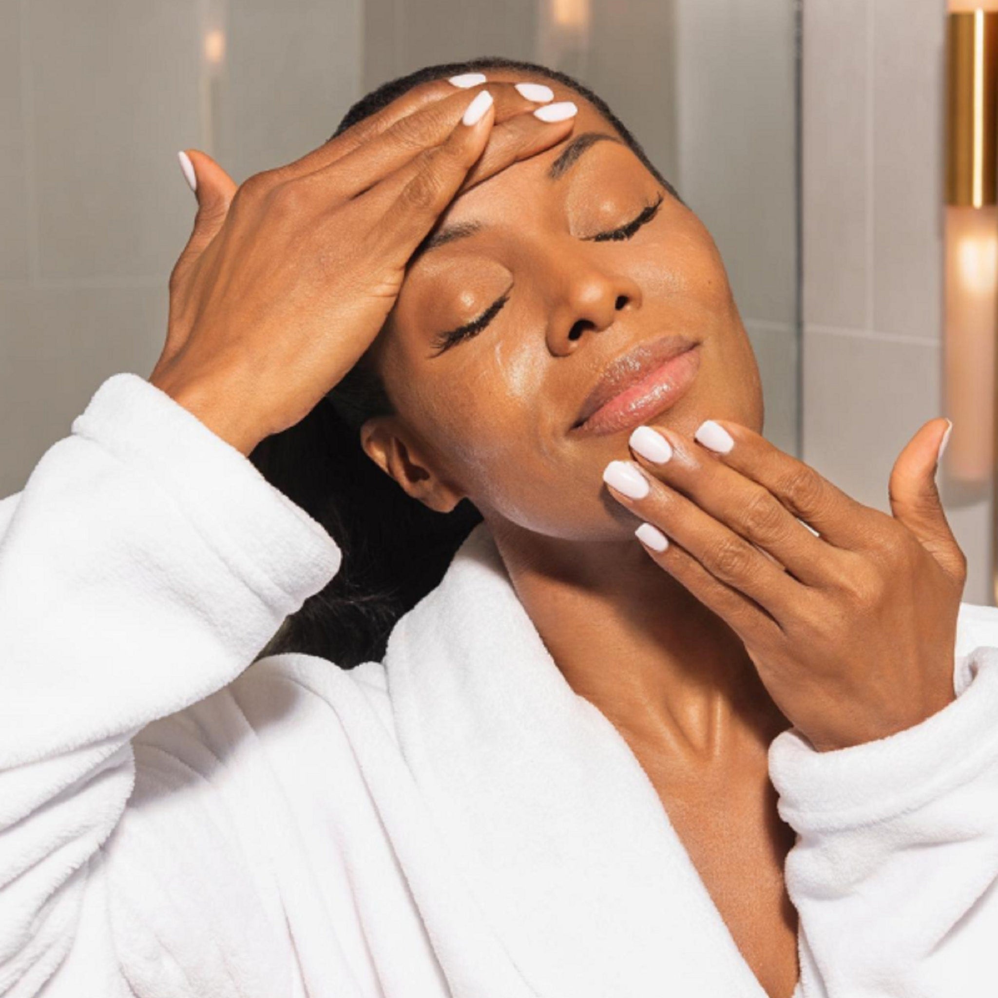 Woman in white robe with closed eyes, applying skincare to face with white nails
