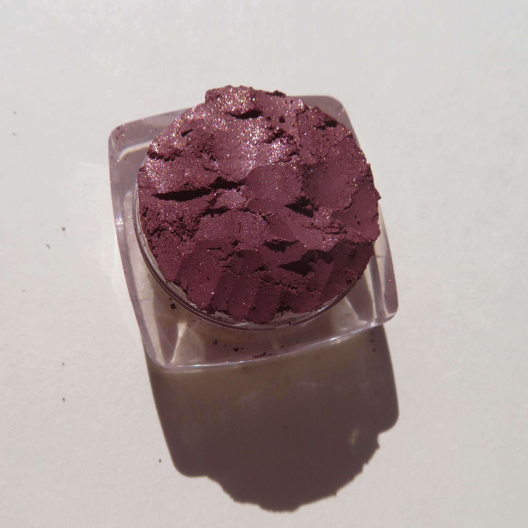 Close-up of loose vegan mineral eye shadow in dark mauve with shimmering pink sheen, showcasing fine pigment texture.