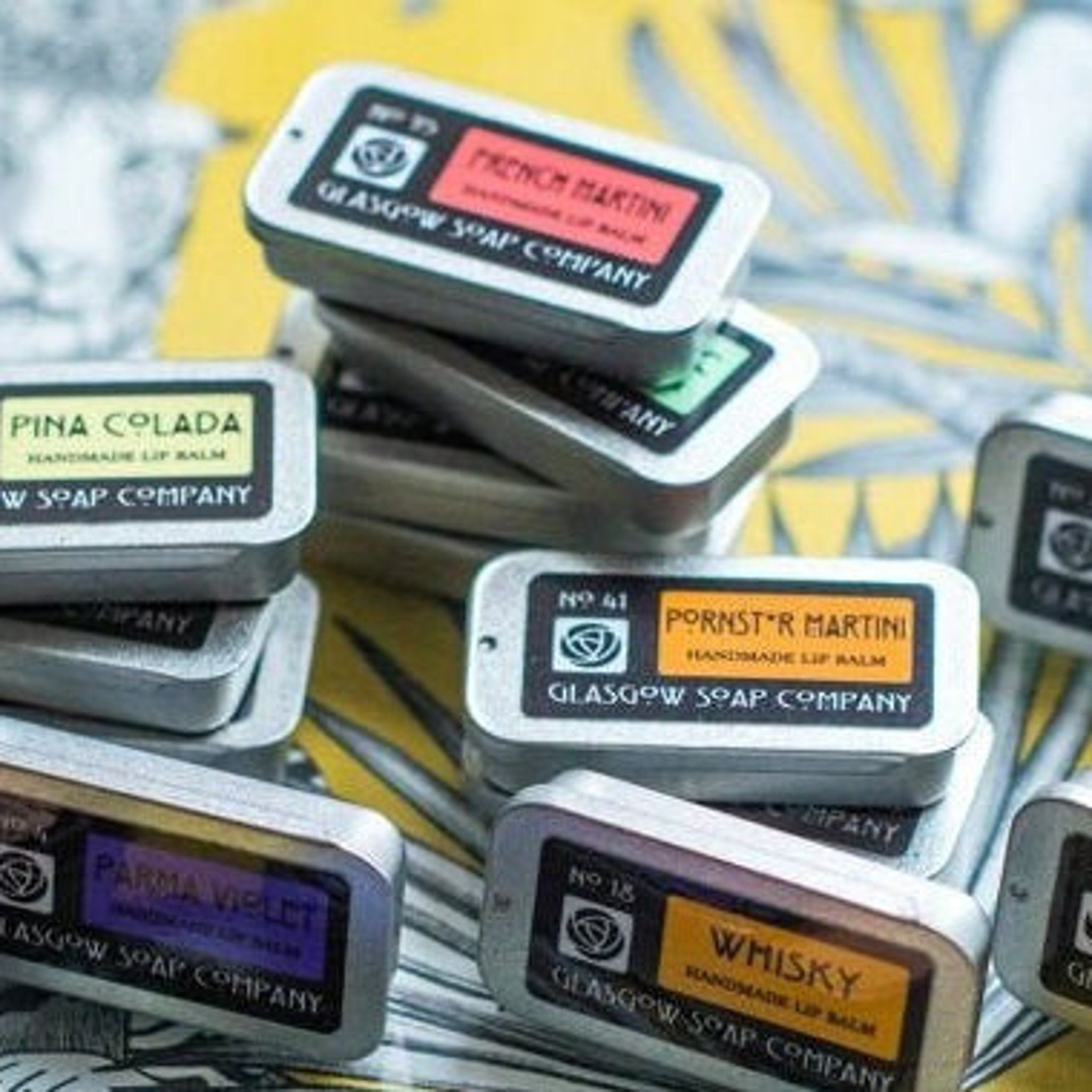 Lip balms in silver tins with colorful labels, Glasgow Soap Company branding visible, stacked together