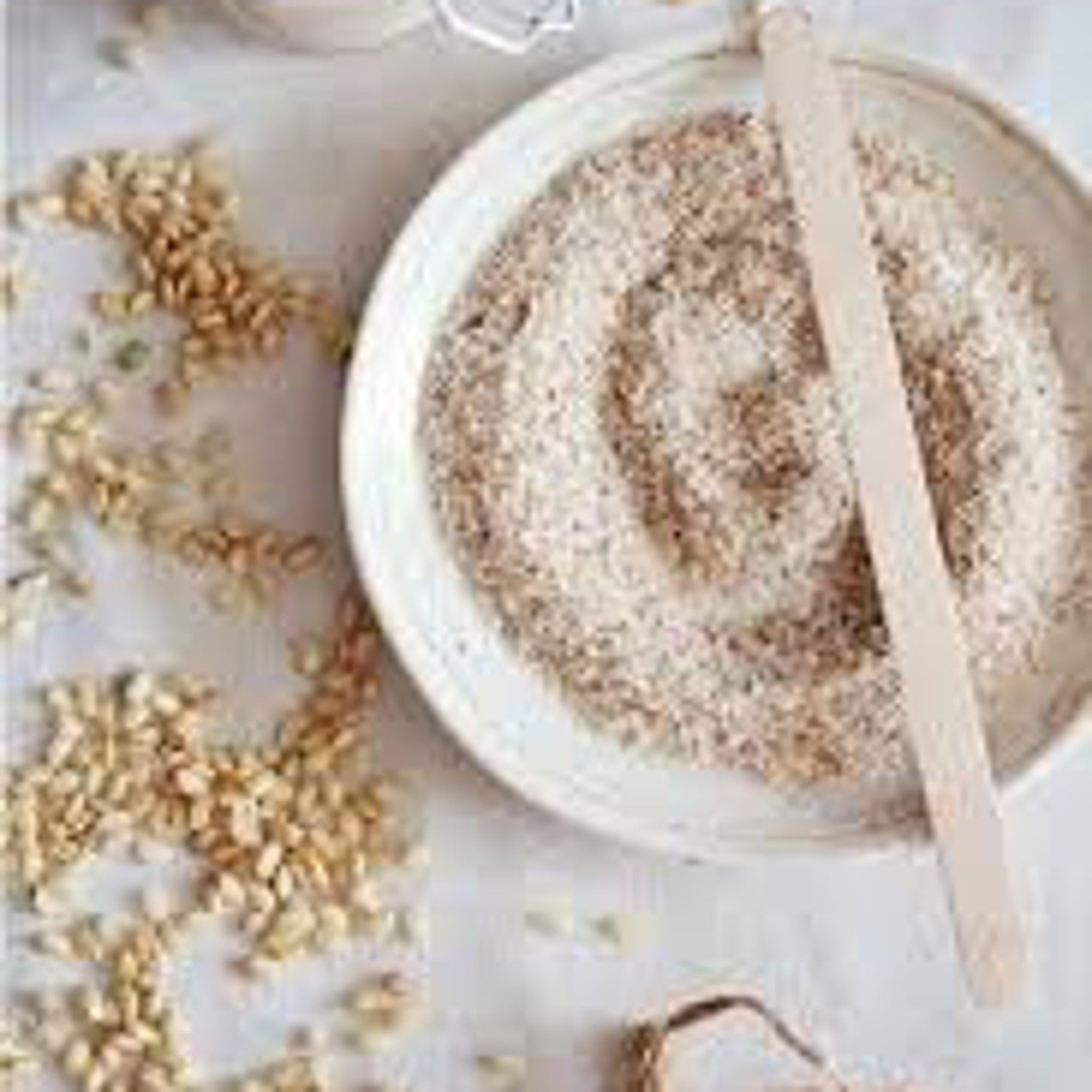 Grain seeds with light brown color and scattered on white surface with bowl of powder and wooden stick