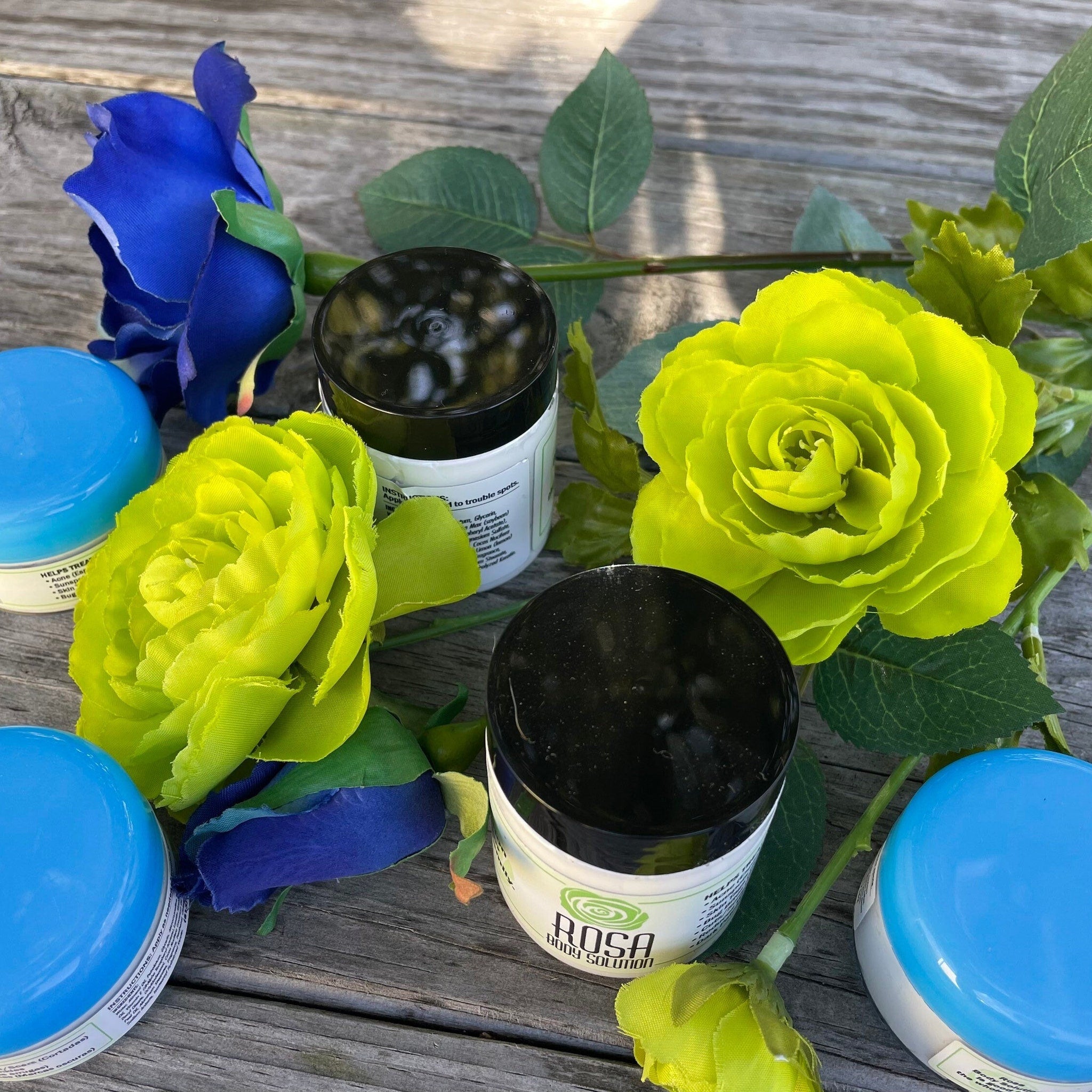 Body cream jars with black lids, blue and yellow flowers, and Rosa Body Solution label on wood surface