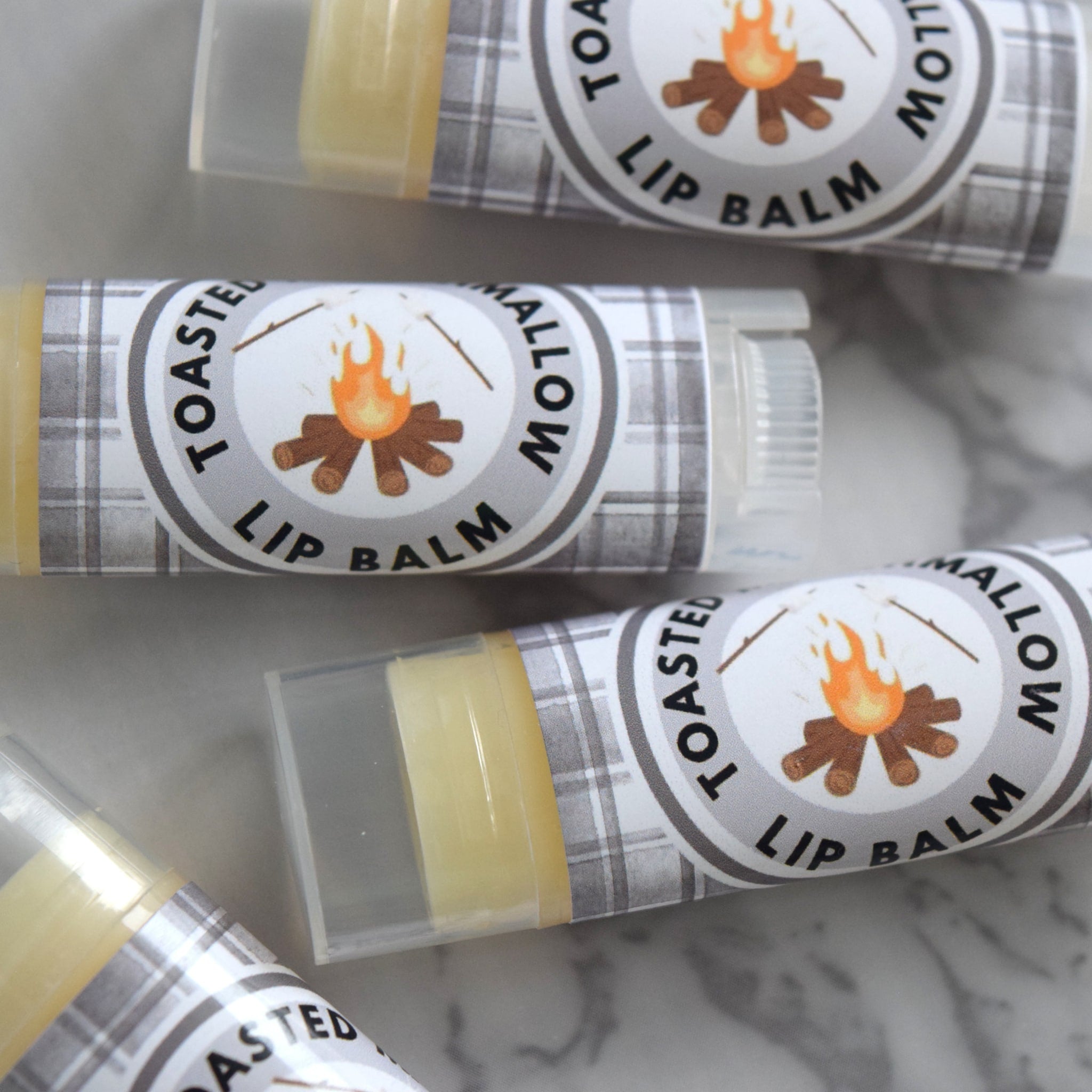 Lip balm tubes with plaid pattern labels, orange and brown campfire graphic, and "Toasted Marshmallow" text