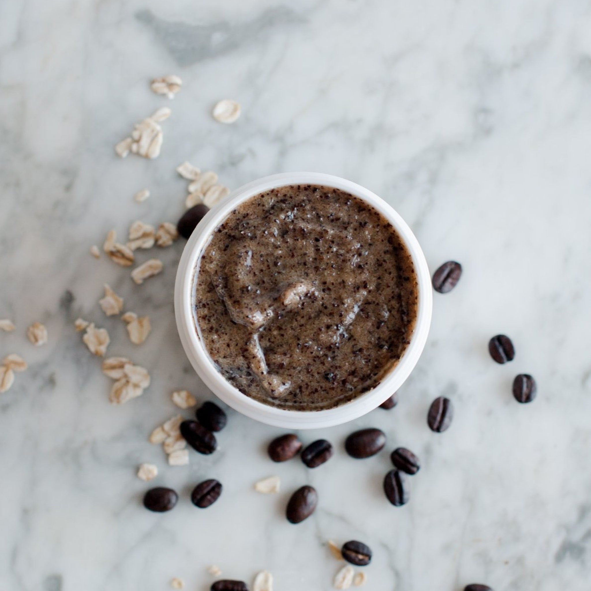 Coffee scrub in white bowl with brown granules and scattered coffee beans and oats