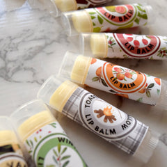 Lip balm tubes with clear plastic caps, various fruit and pattern labels, and yellow balm inside