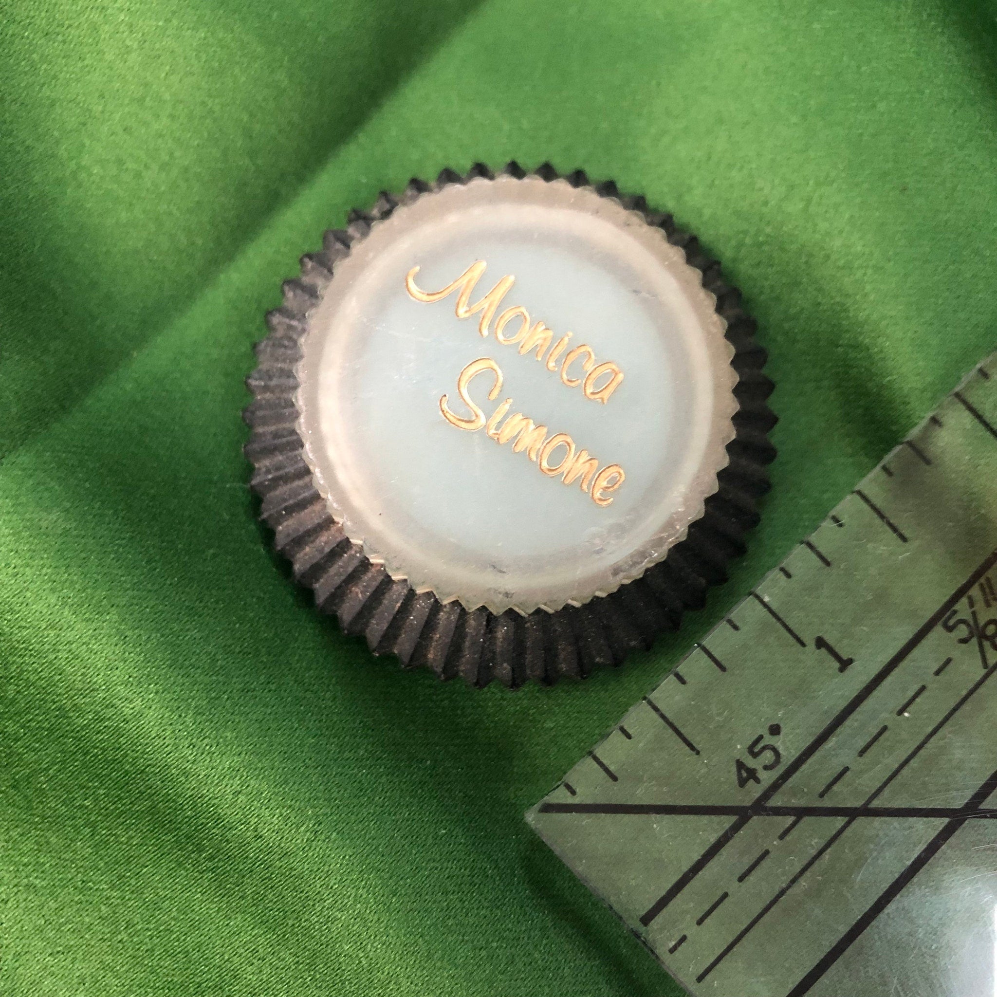Round name tag with gold text, black ridged edge, and light blue center on green fabric
