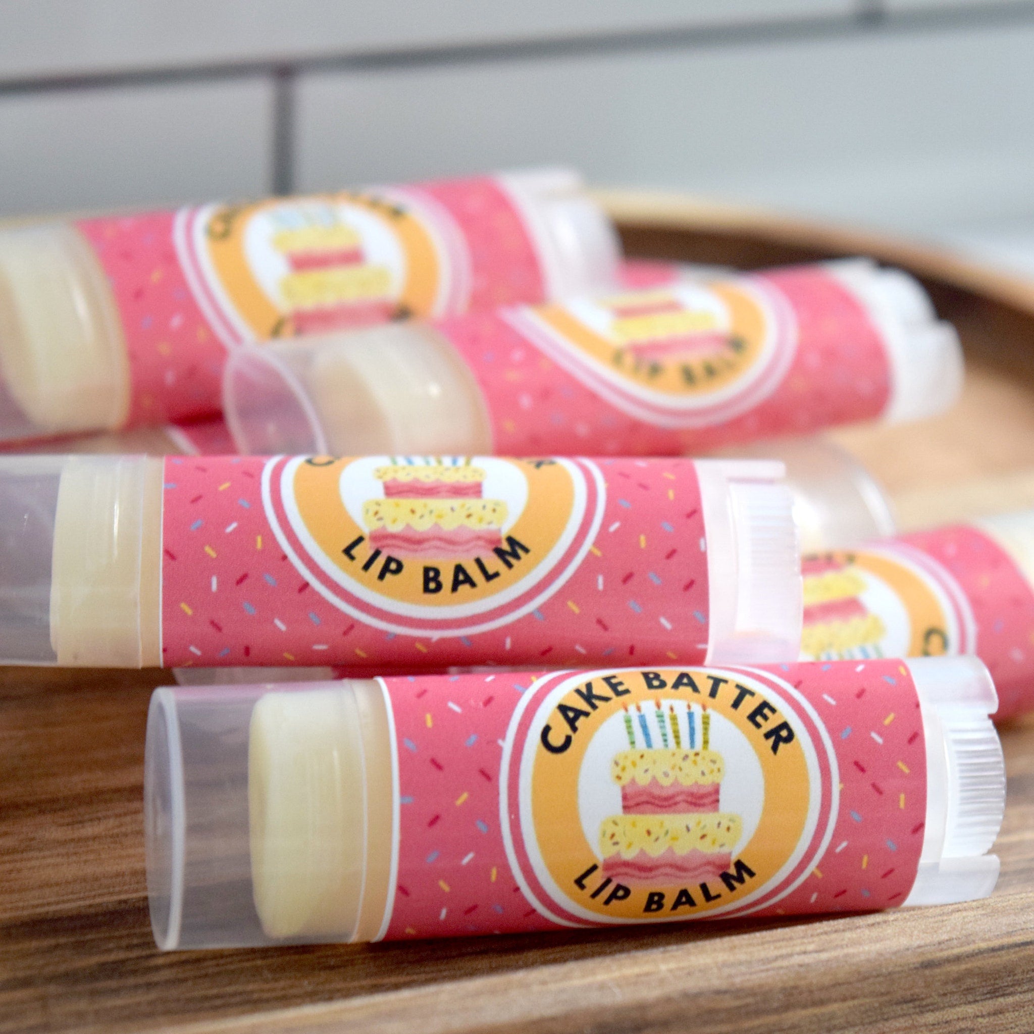 Lip balm tubes with pink labels, colorful sprinkles, and cake design on wooden surface