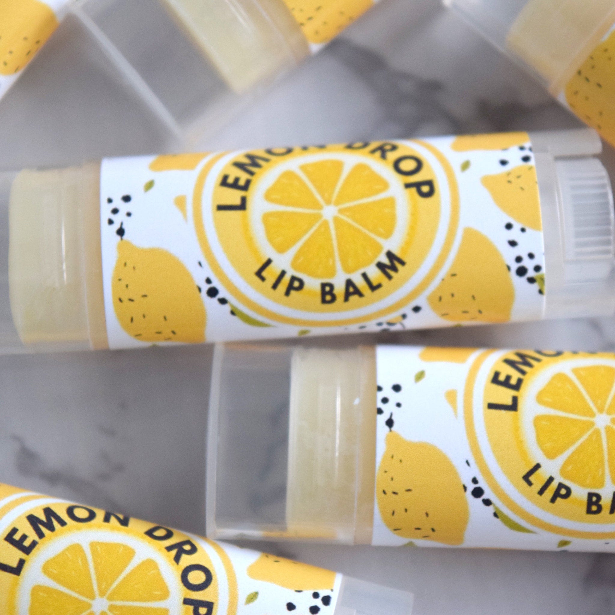 Lip balm tubes with yellow and white "Lemon Drop" label, clear plastic container, and yellow balm inside