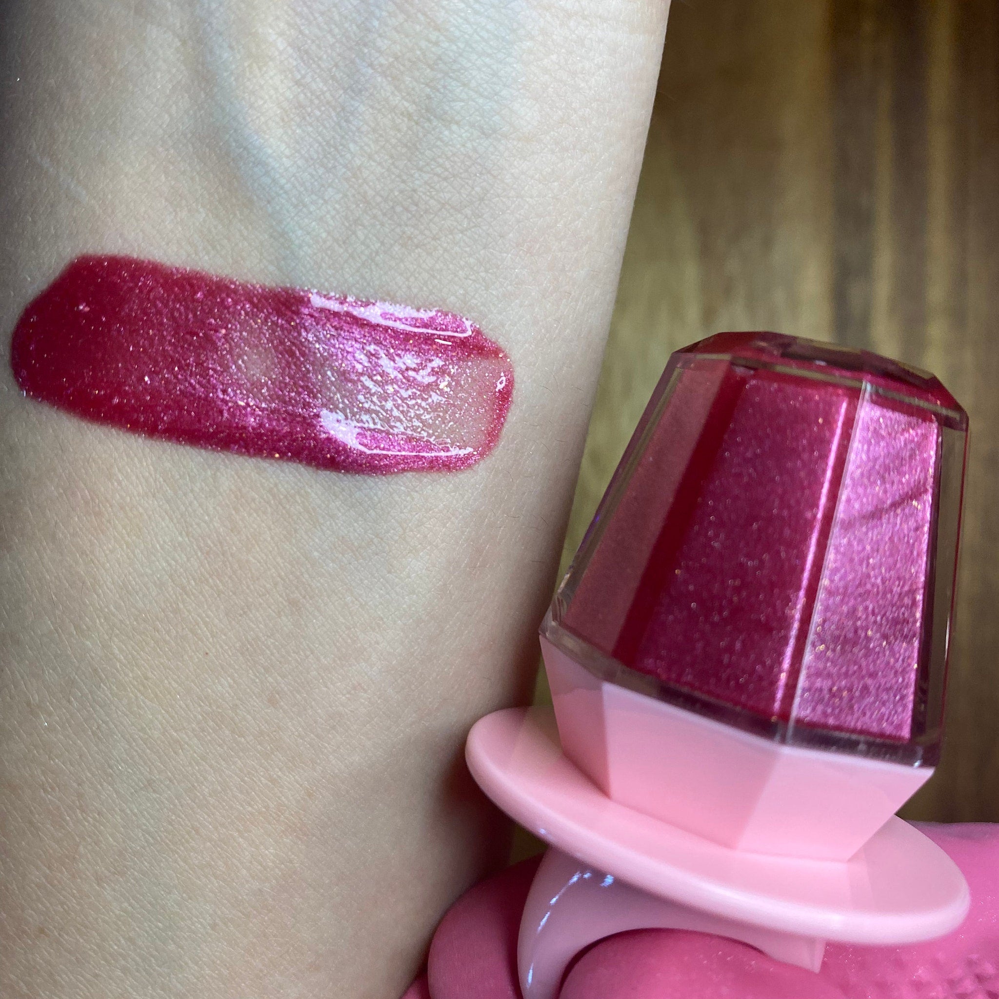Lip gloss swatch with pink shimmer and diamond-shaped container on skin, showing color and texture