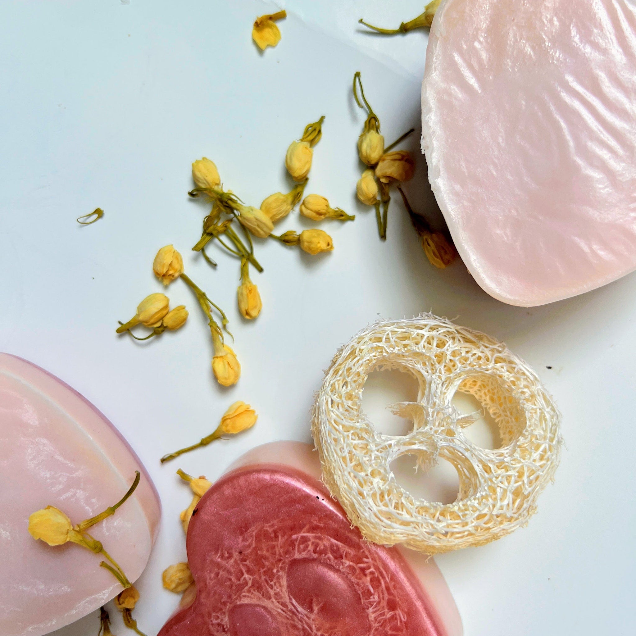 Soaps with pink and red hues, loofah, and dried yellow flowers on white background