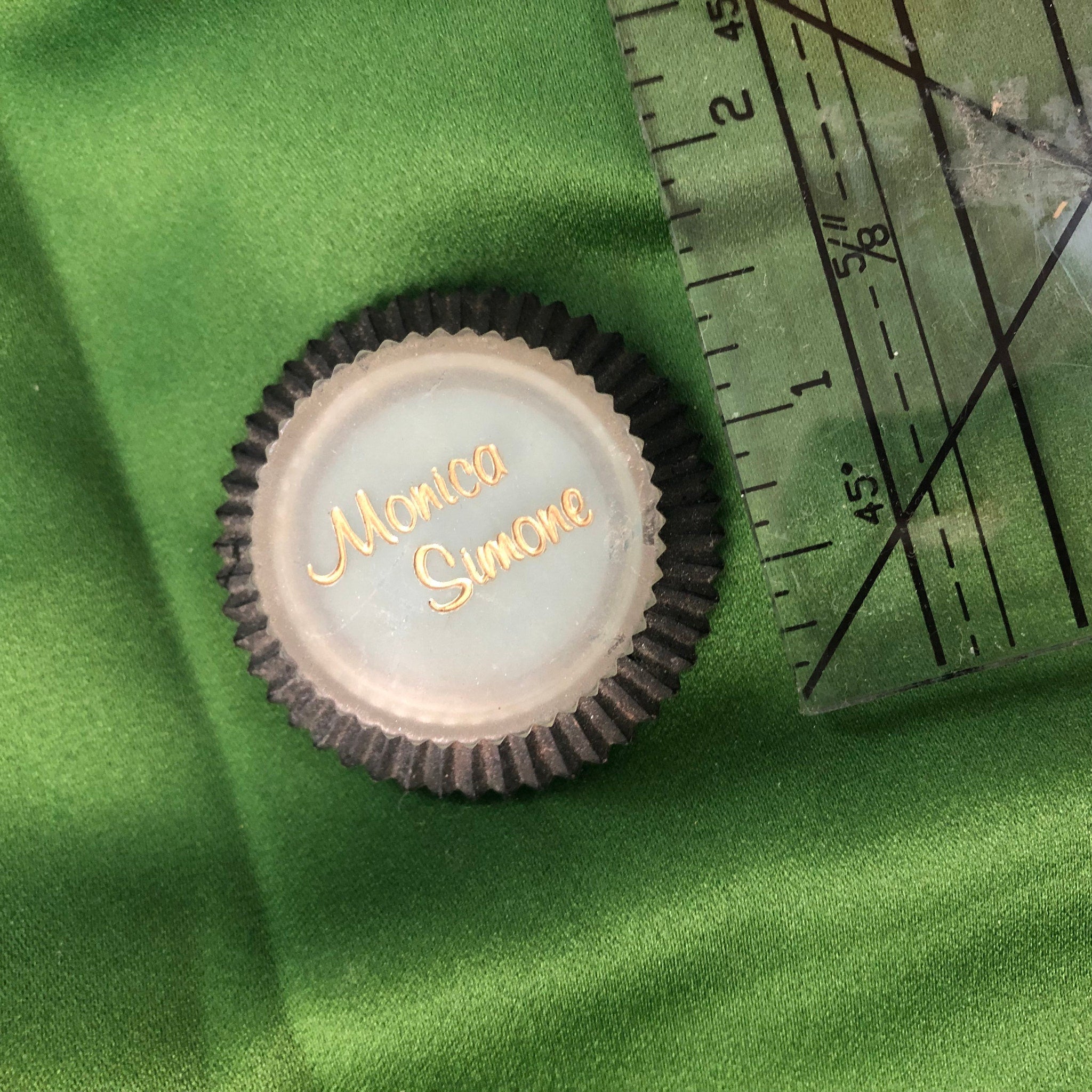 Round name tag with gold lettering, black edge, and green fabric background with ruler