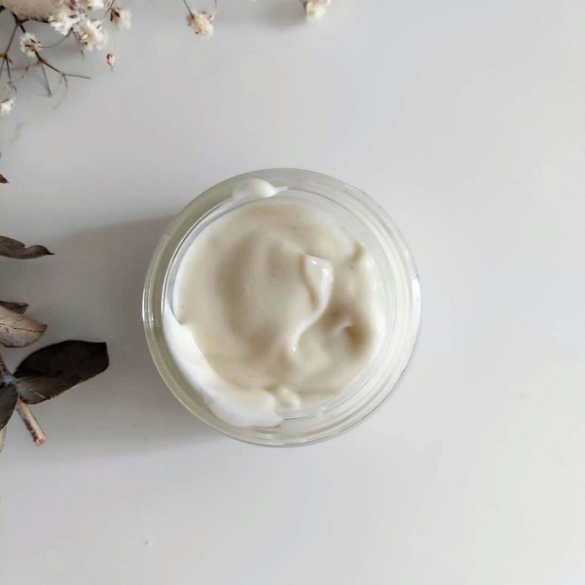 Cream jar with white cream, clear glass, and plant sprig on white background