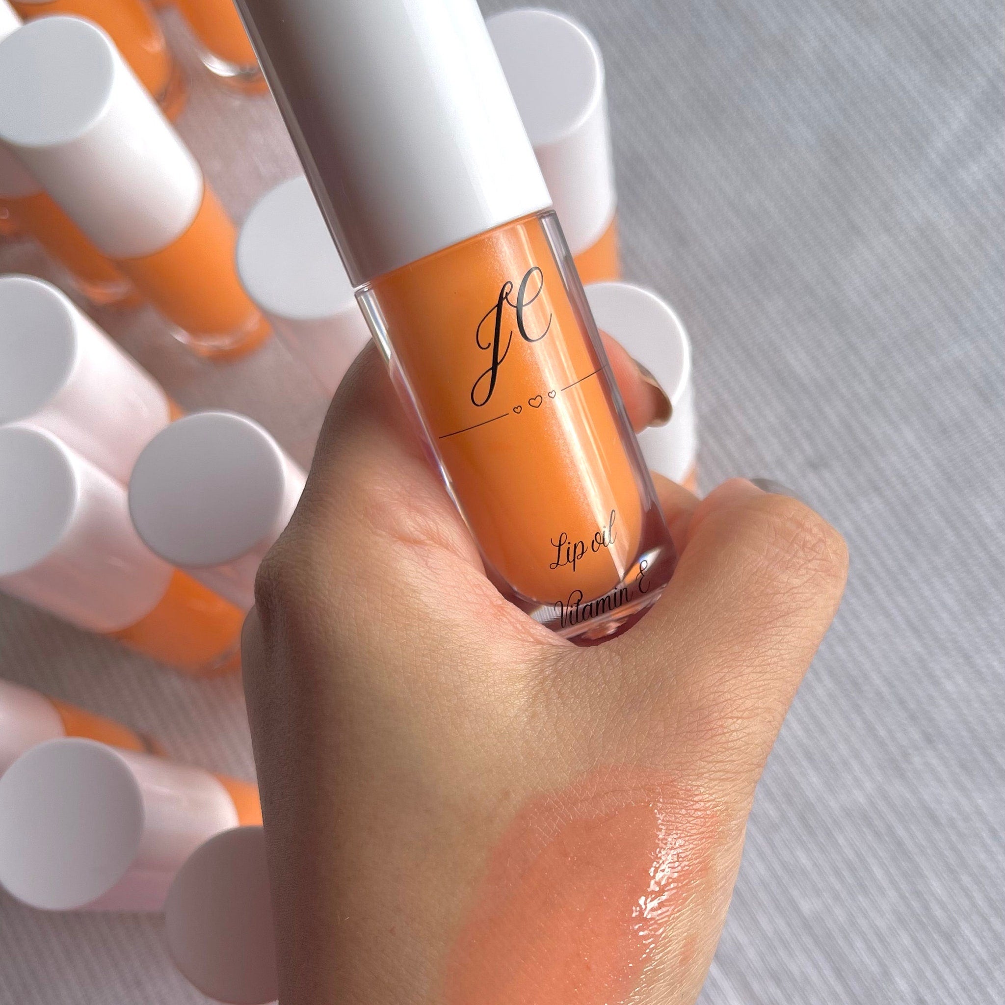 Lip oil with orange tint, clear bottle, white cap, and "JC" logo, held in hand