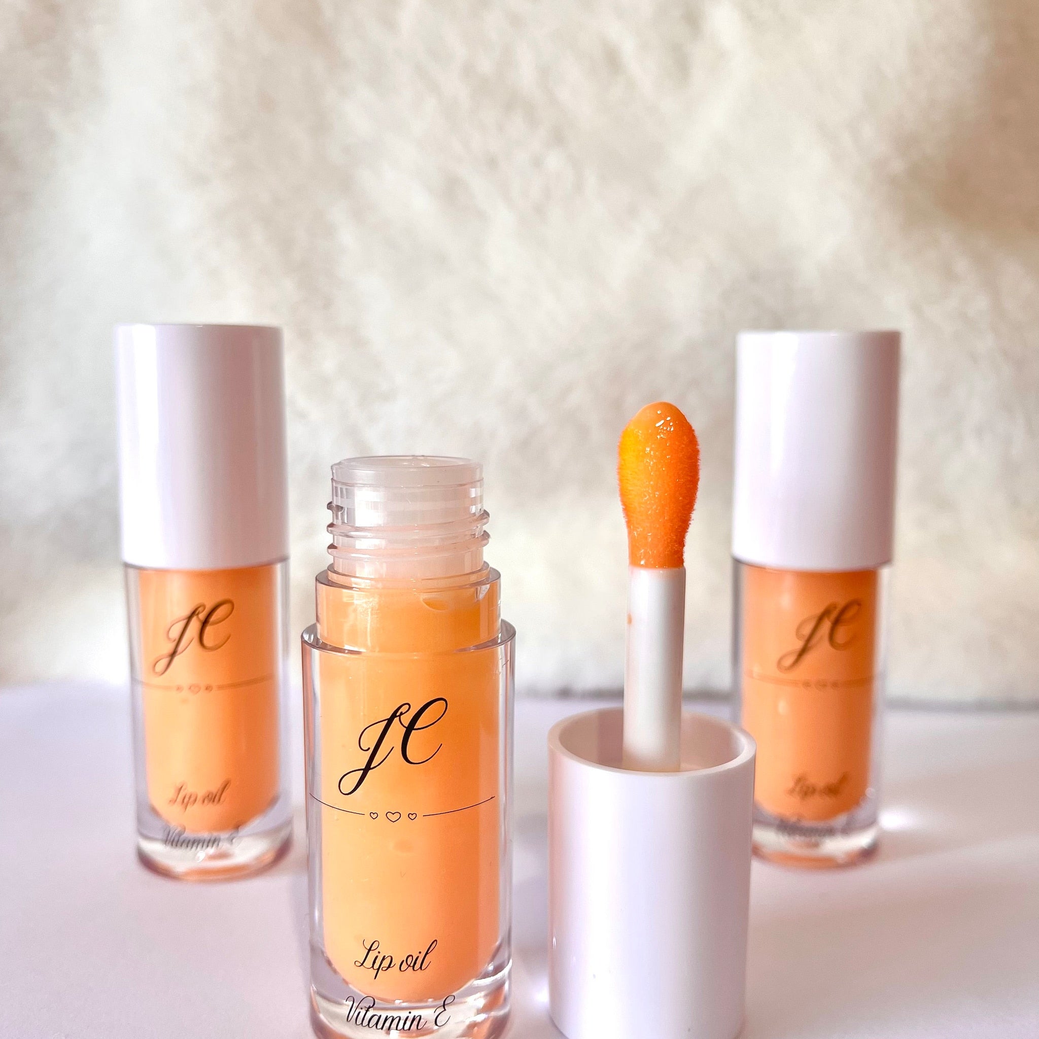 Lip oil bottles with orange liquid, white caps, and JC text, Vitamin E label visible