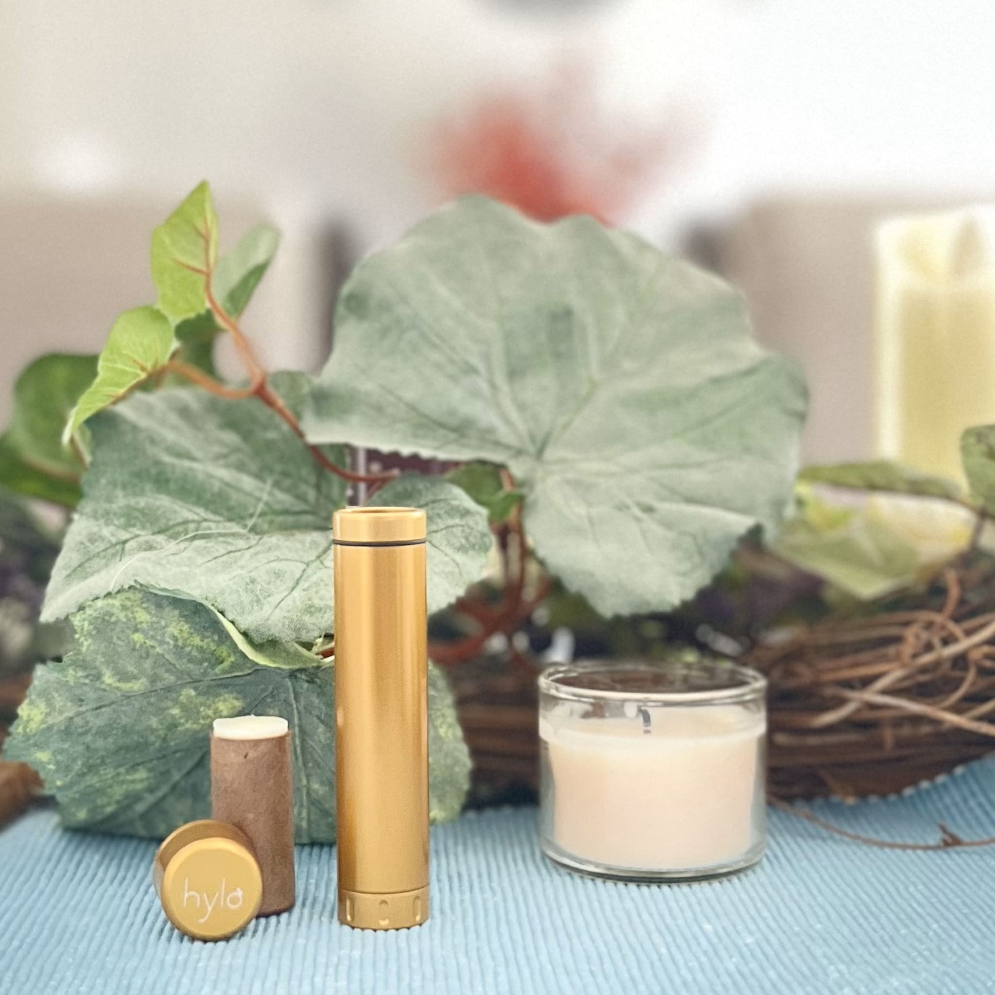 Gold metal tumbler with hylo logo, brown candle, and lit white candle on blue surface