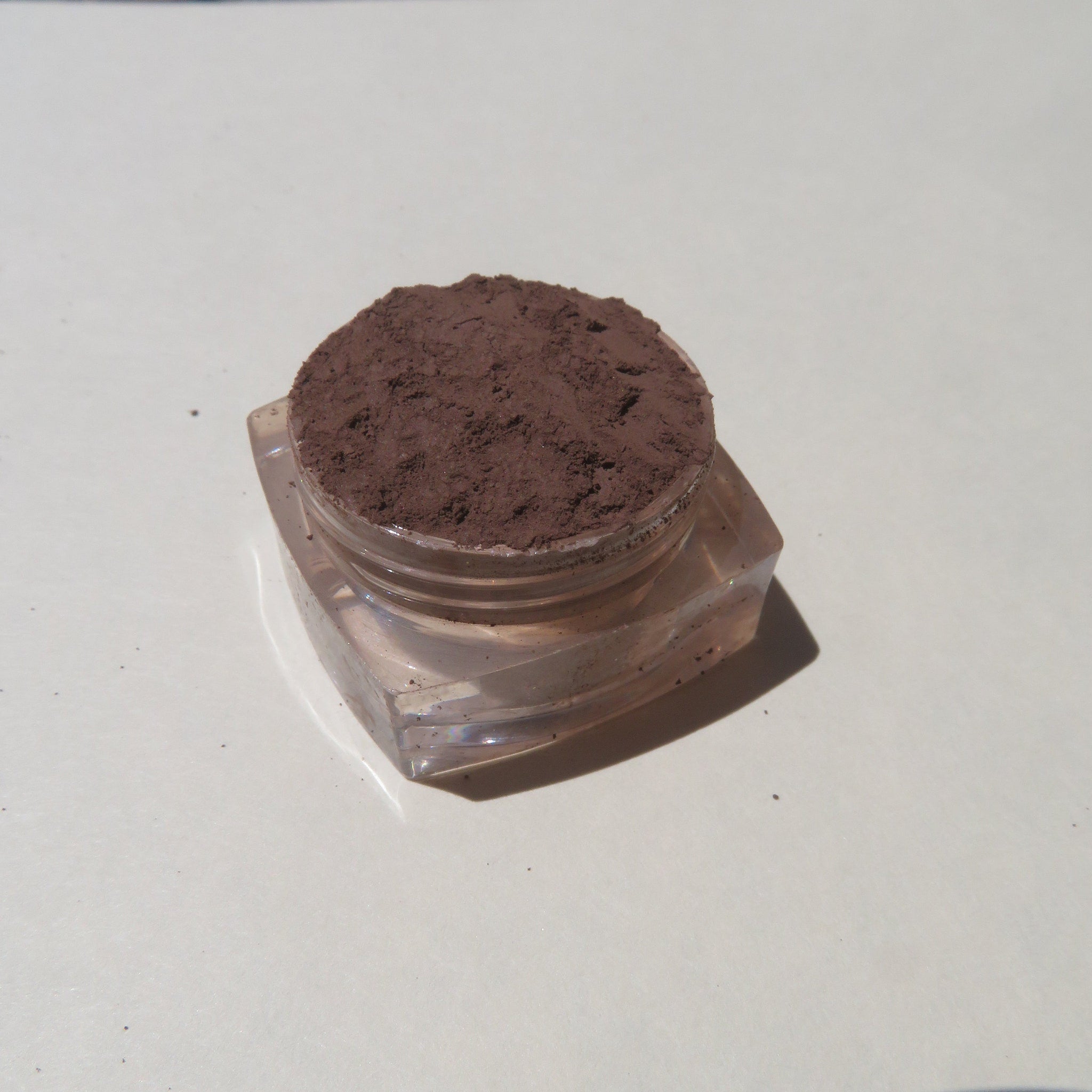 Close-up of matte dark brown mineral eyeshadow powder spilling from jar, showcasing rich pigment texture