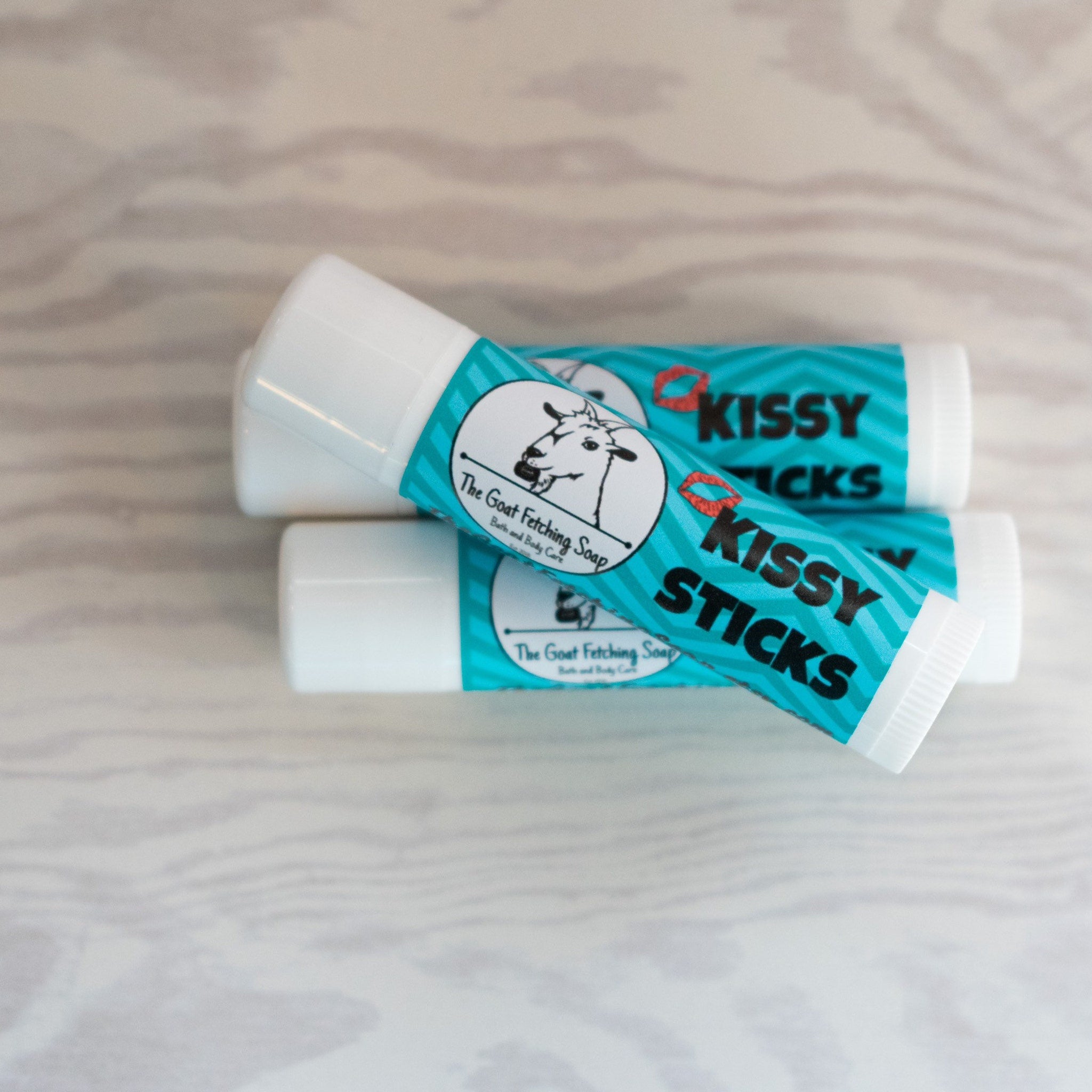 Lip balm tubes, white and teal, with "Kissy Sticks" text and Goat Fetching Soap logo on each tube