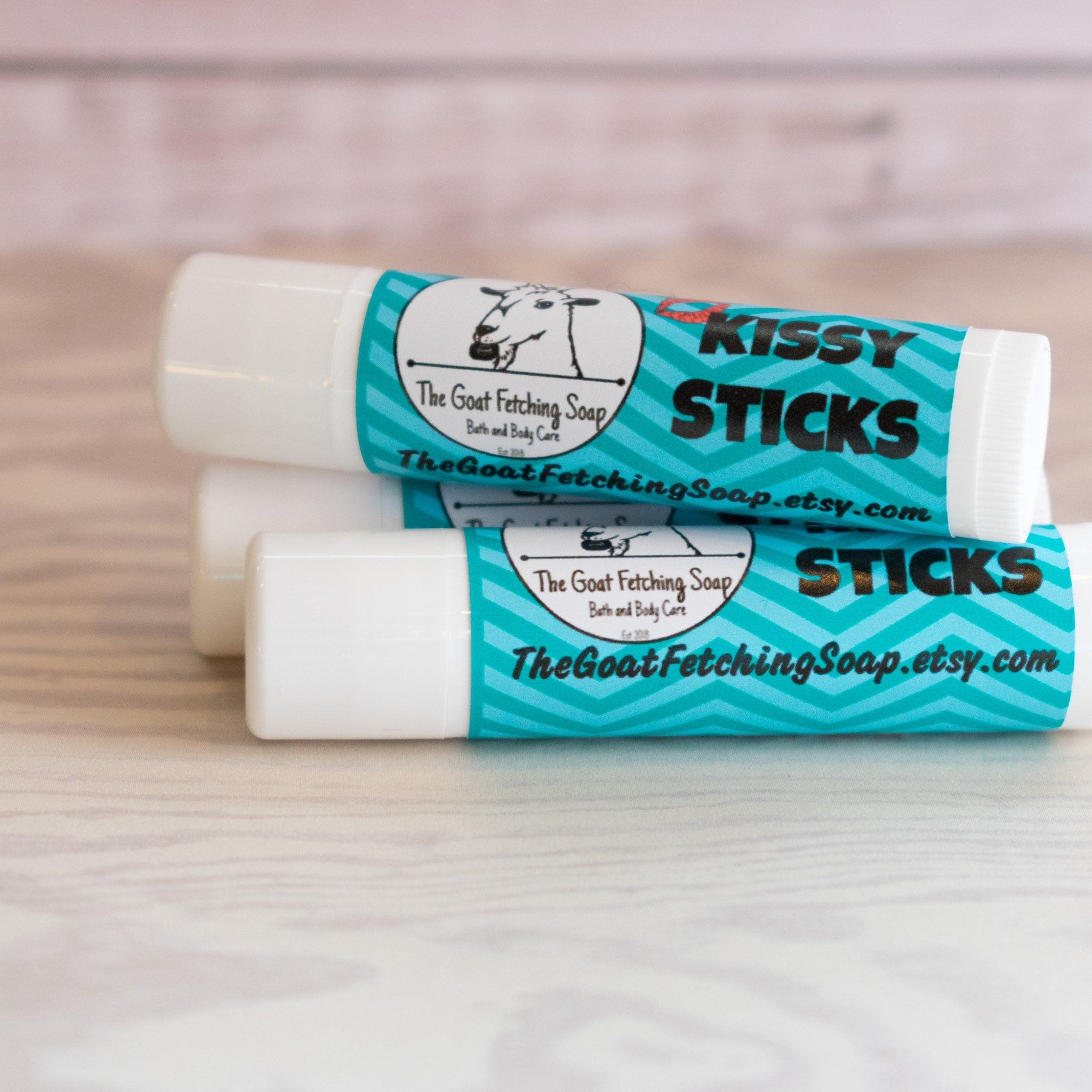 Lip balm tubes with white and teal chevron labels, "Kissy Sticks" text, and The Goat Fetching Soap brand