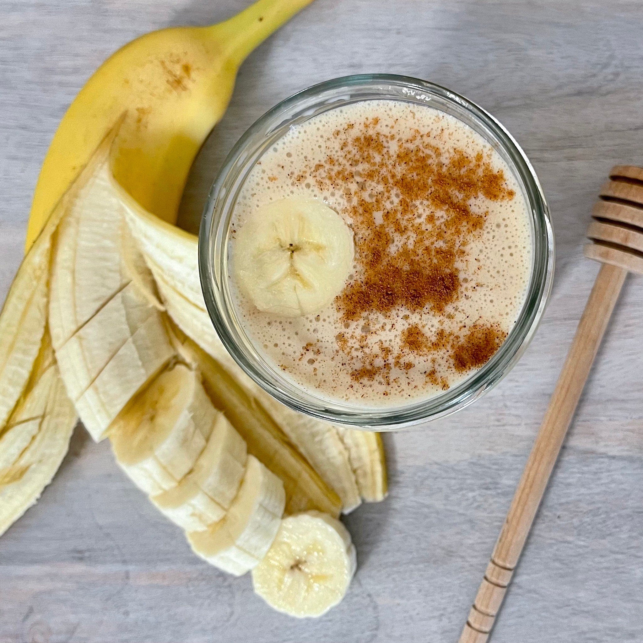 Banana smoothie in glass with sliced banana, brown spice, and wooden honey dipper on a gray surface