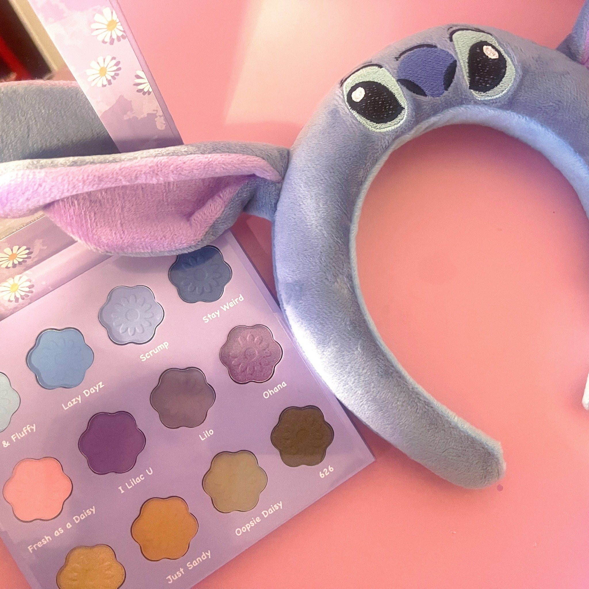 Makeup palette with flower-shaped eyeshadows in purple, pink, and blue shades, with a Stitch headband on a pink surface