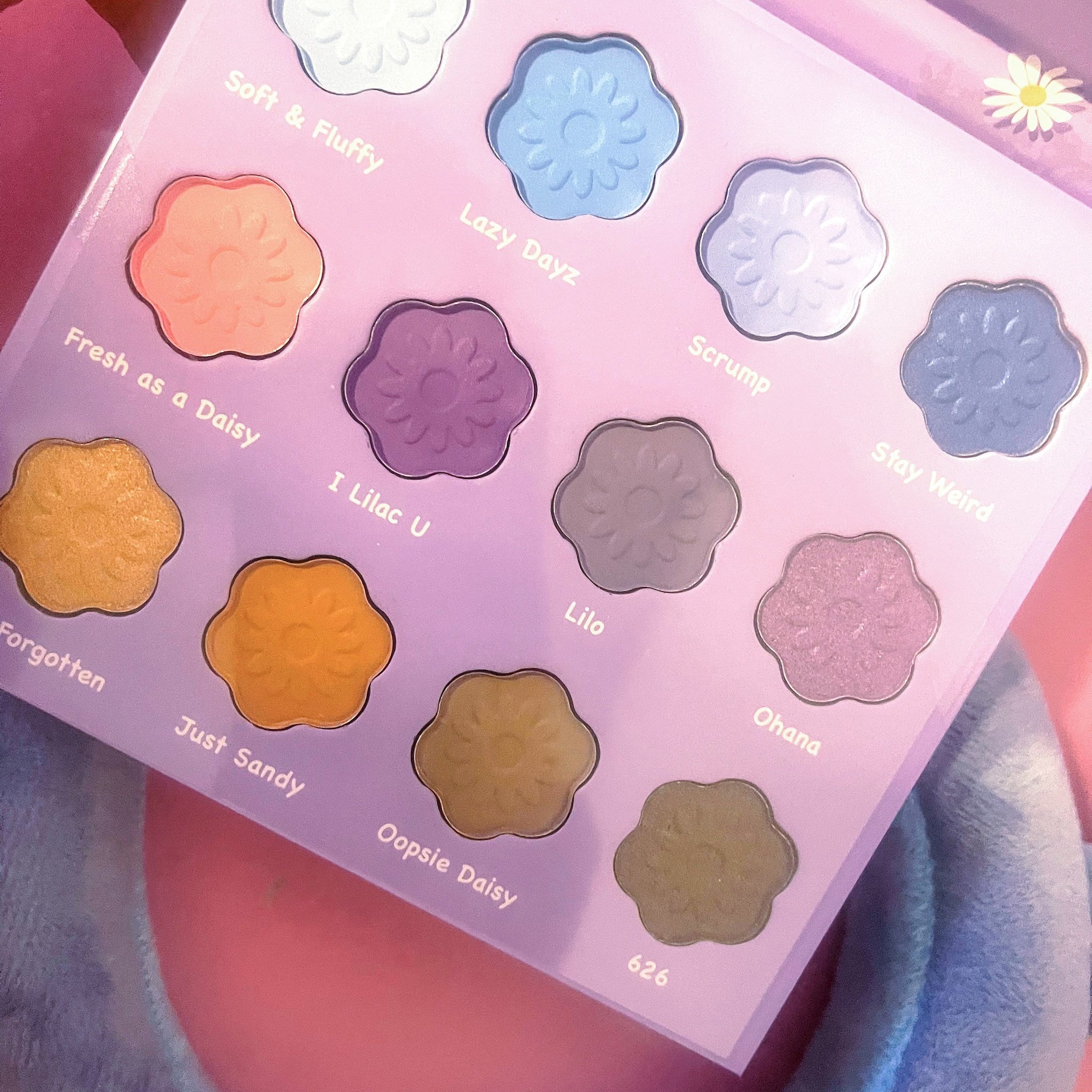 Eyeshadow palette with flower-shaped pans in various colors on a lavender background, names visible