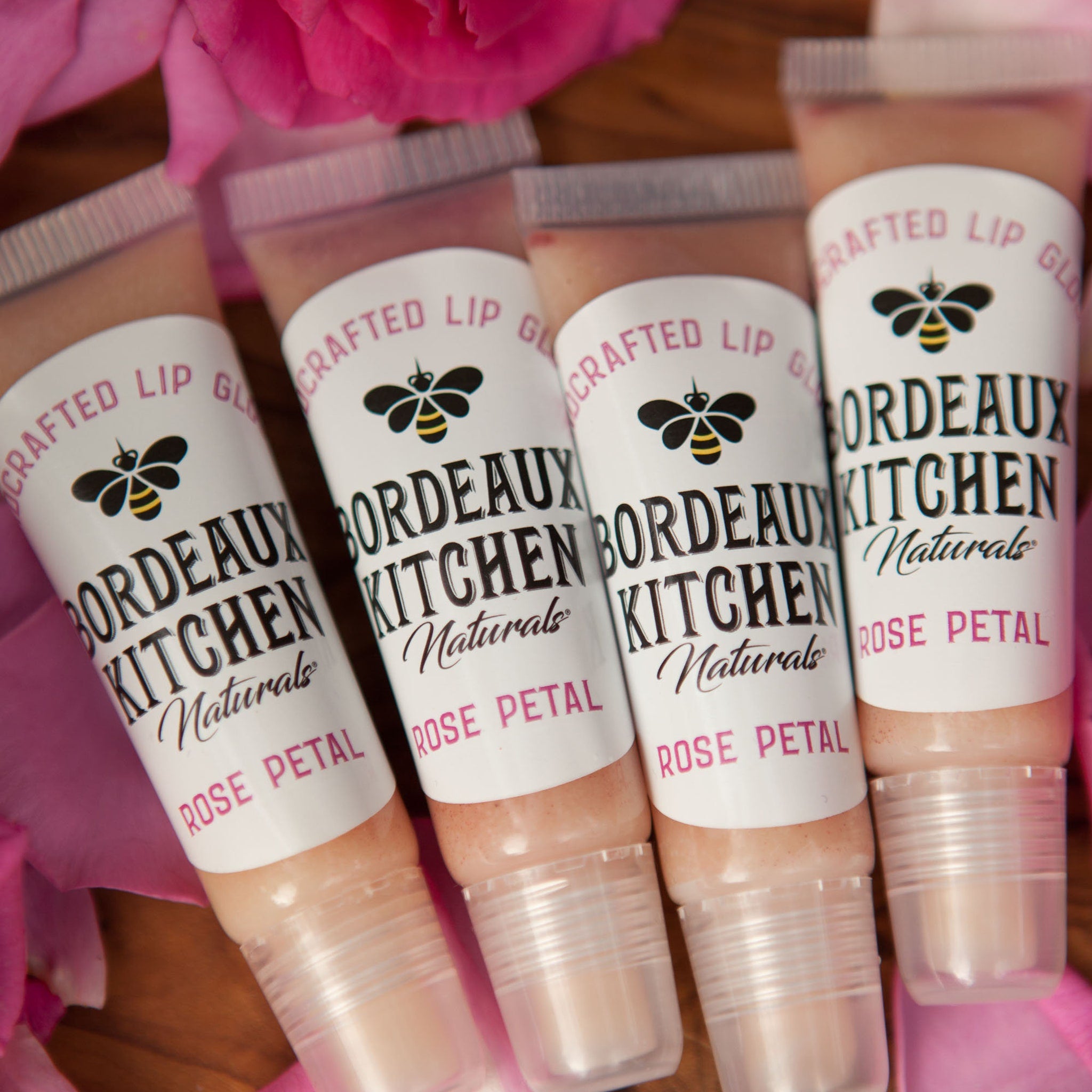 Lip gloss tubes with pink liquid, white labels, and "Bordeaux Kitchen Naturals" text, surrounded by pink rose petals
