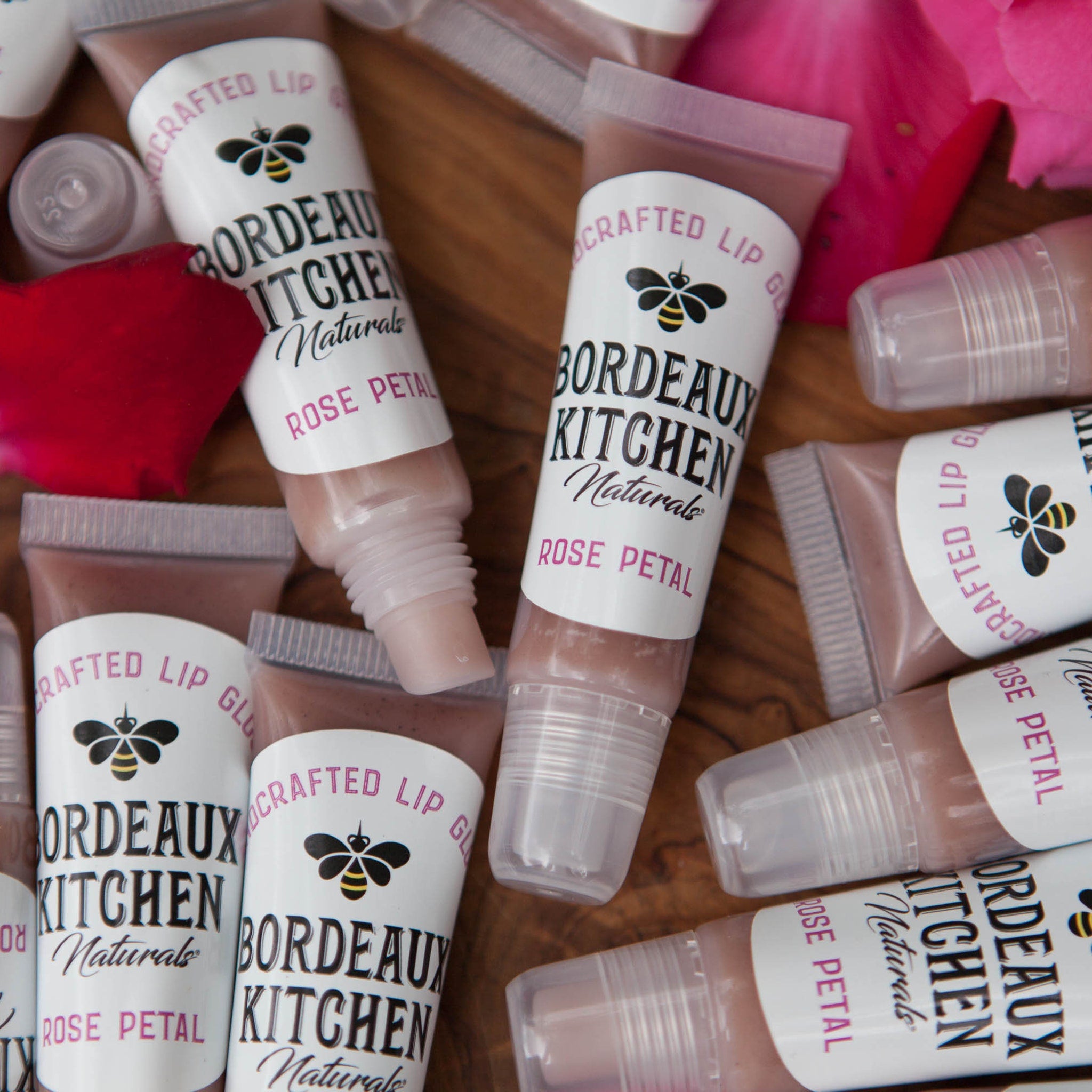 Lip gloss tubes with clear plastic, pink gloss, and Bordeaux Kitchen label on wood surface