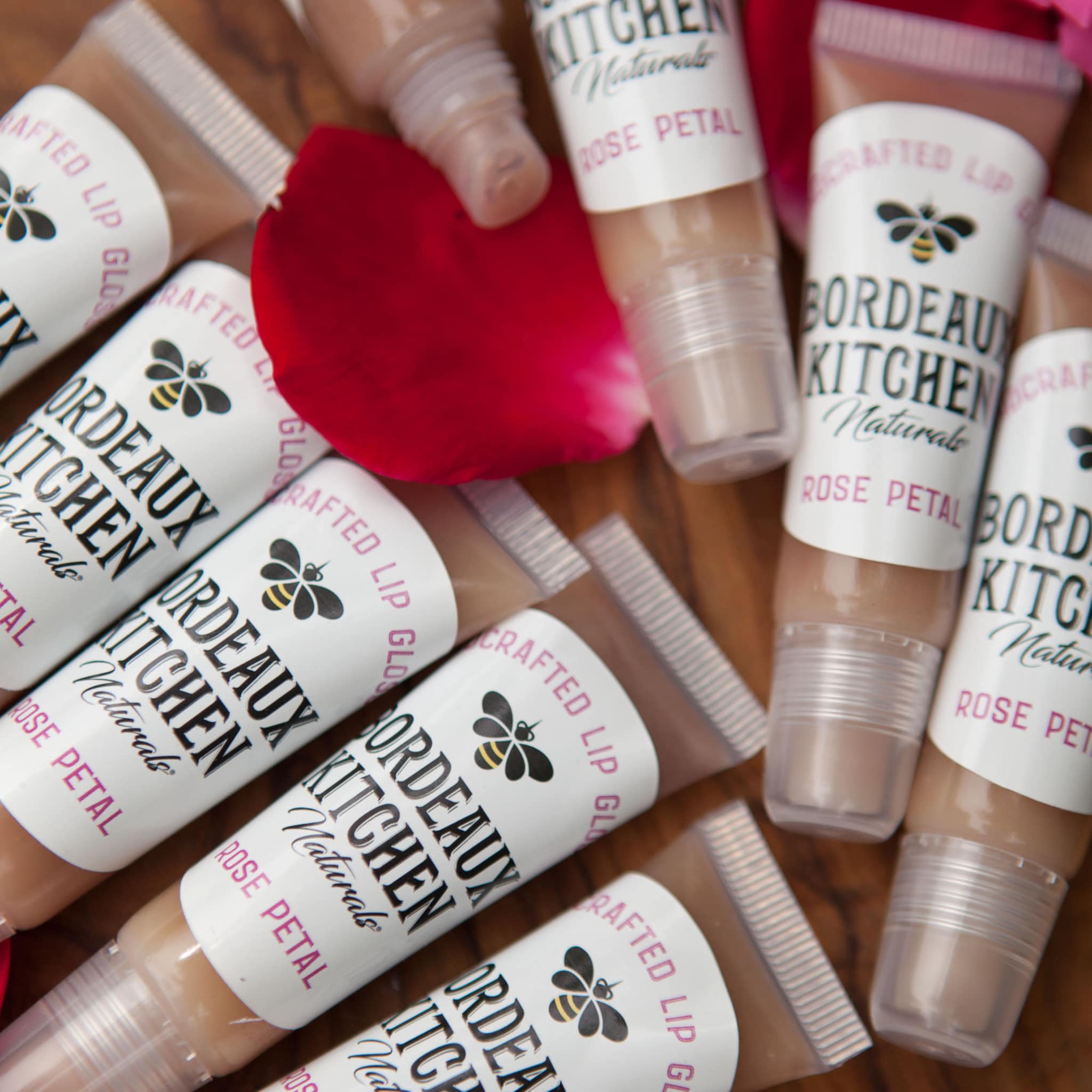 Lip gloss tubes with clear plastic, beige gloss, and Bordeaux Kitchen label on wooden surface