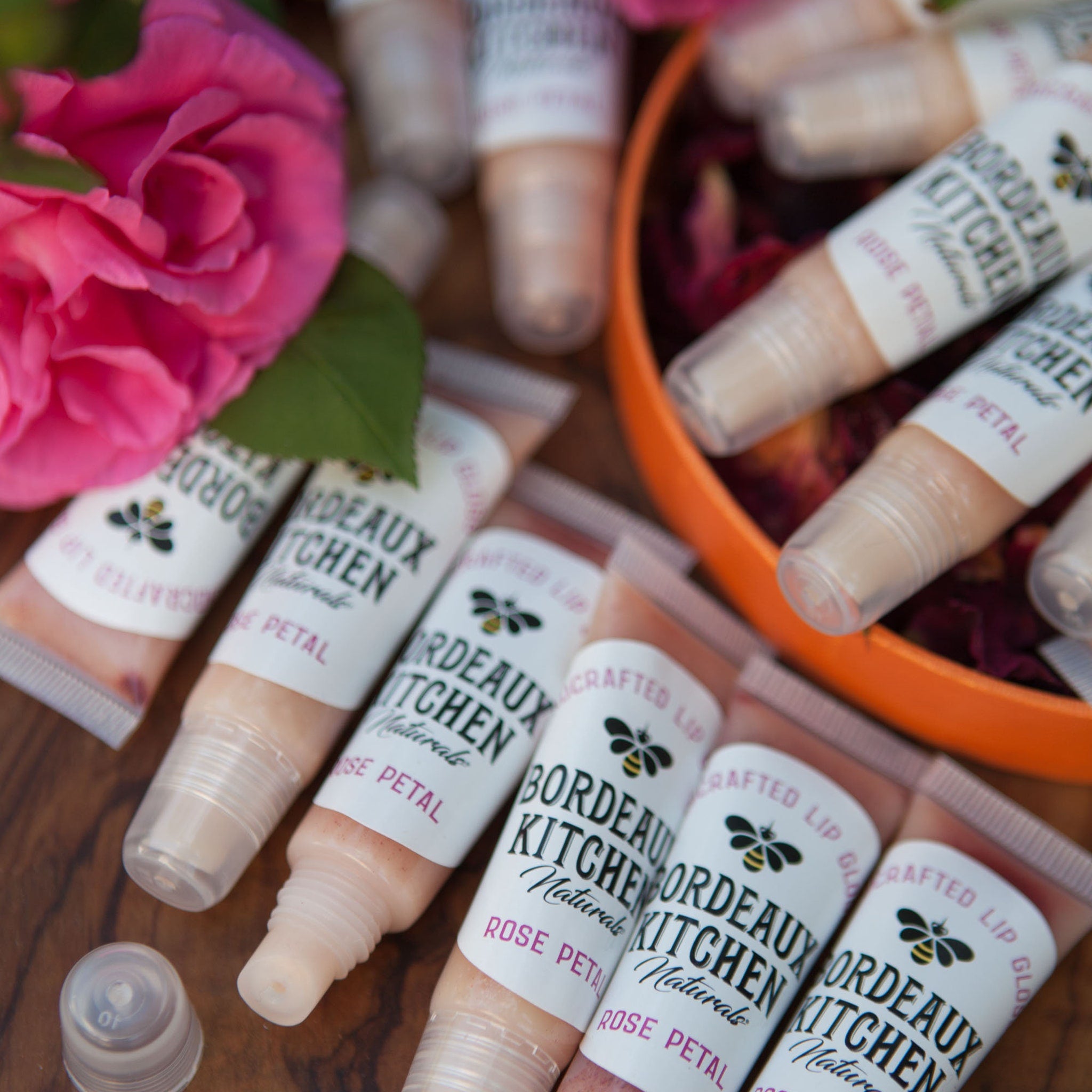 Lip gloss tubes with pink tinted gloss, white labels, and Bordeaux Kitchen branding, arranged with roses