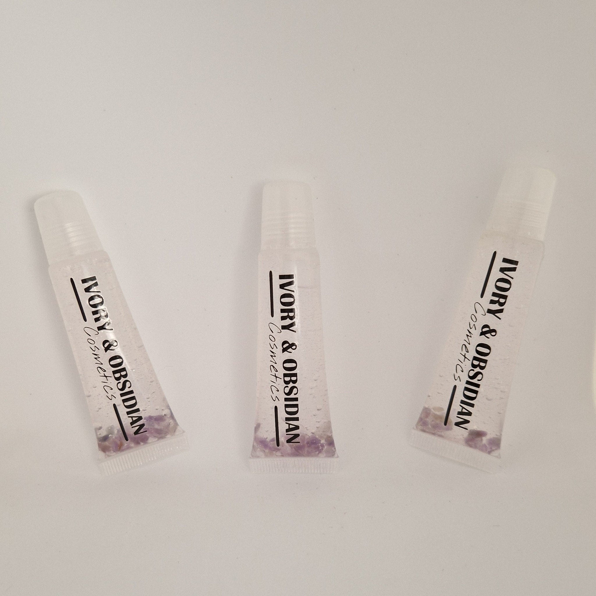 Lip gloss tubes with clear liquid, purple stones, and Ivory & Obsidian Cosmetics text on white background