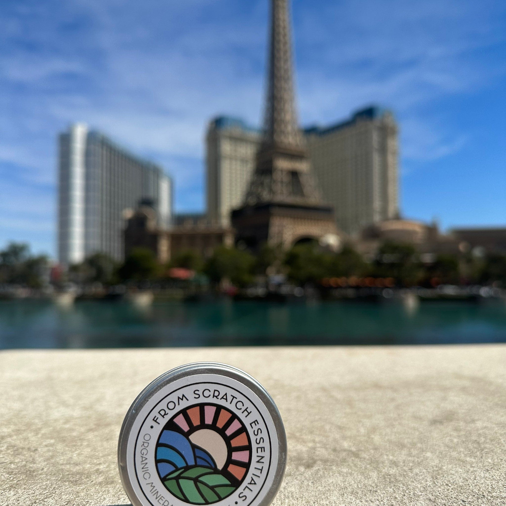 Round metal tin with white label, colorful logo, and "From Scratch Essentials" text, outdoors with blurred background