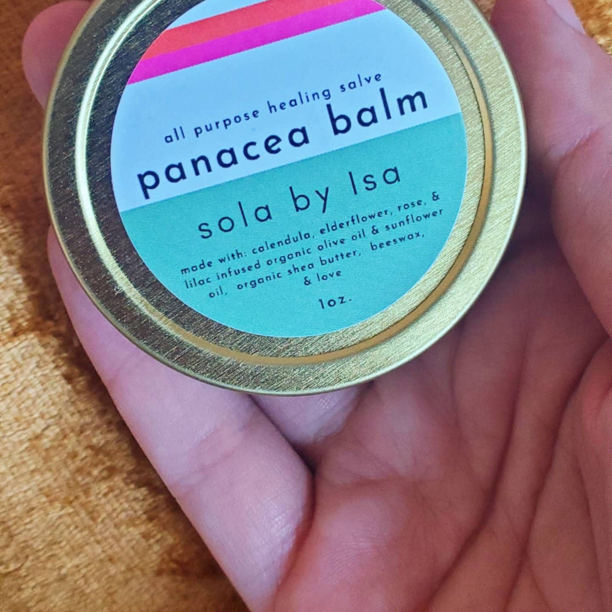 Panacea balm in round metal tin with sola by Isa text, held in hand, teal and gold colors