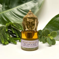 Lip balm jar with gold lid, purple label, and Wise Healer text, surrounded by green leaves