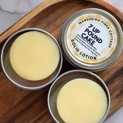 Solid lotion in silver tins with yellow cake, on a wooden tray with "Wandering Pines Cottage" label