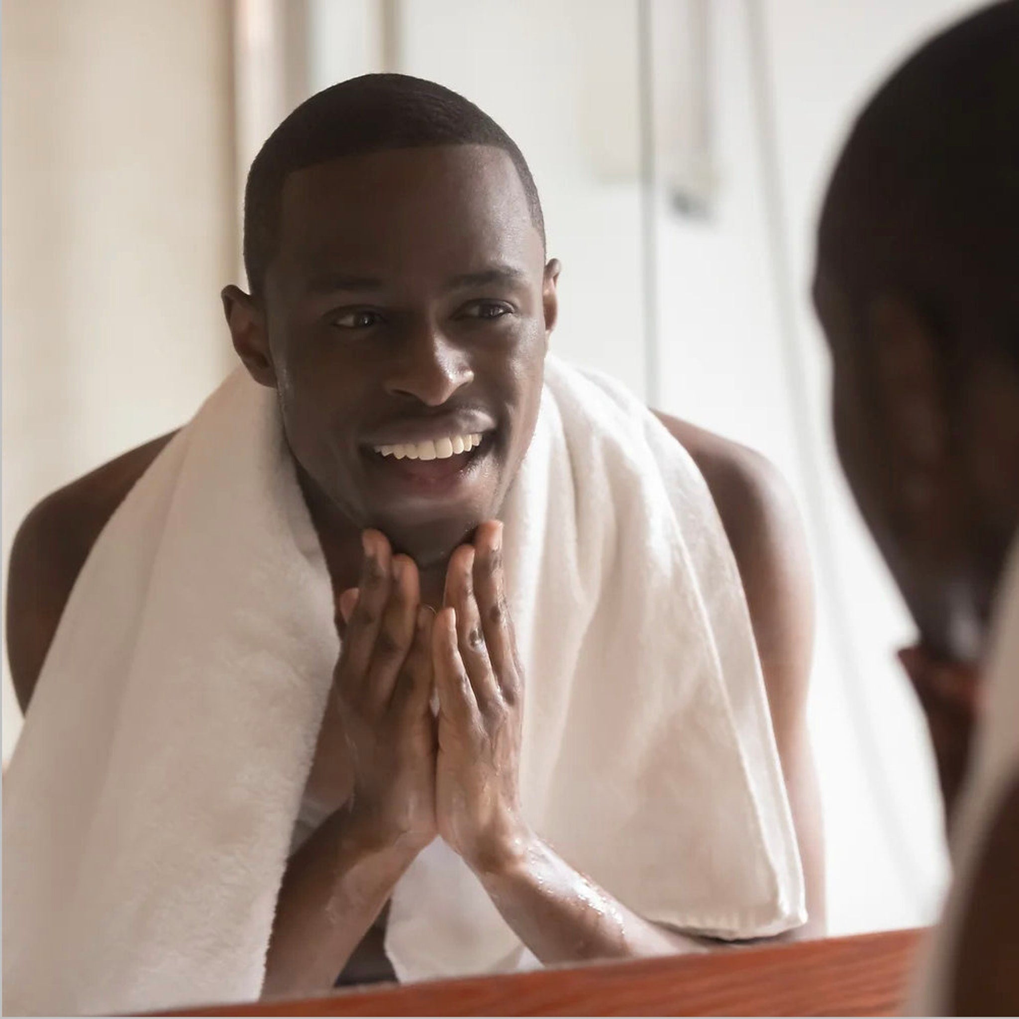 Man with dark hair and skin, smiling, wearing white towel around neck, in front of mirror