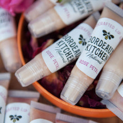 Lip gloss tubes with white labels, clear caps, and "Bordeaux Kitchen Naturals" text, in orange bowl
