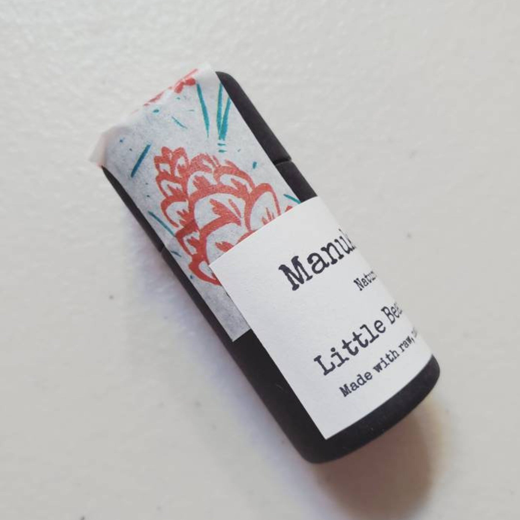Black soap bar wrapped in patterned paper with Manuka text visible