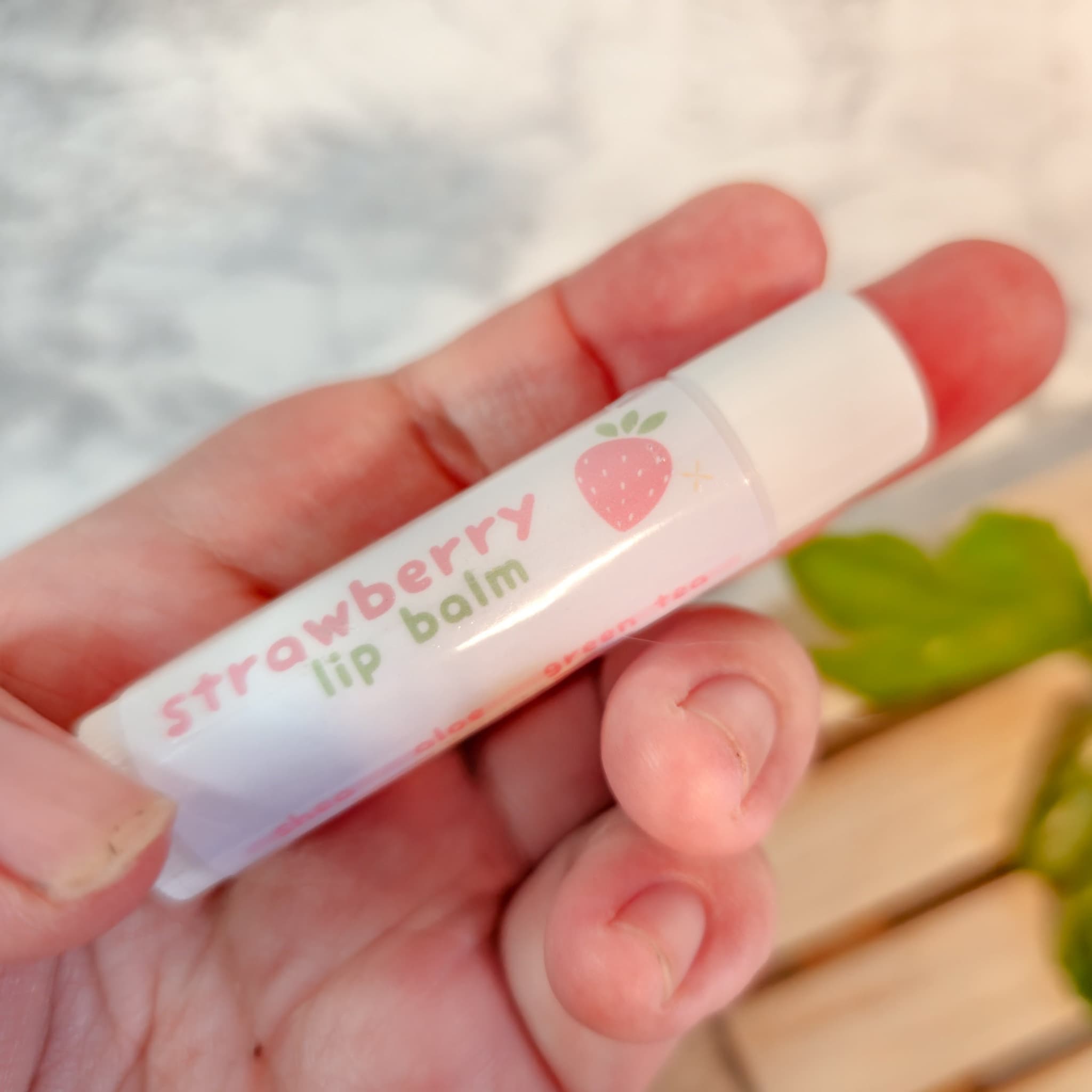 Strawberry lip balm in white tube with pink text and strawberry graphic, held in hand