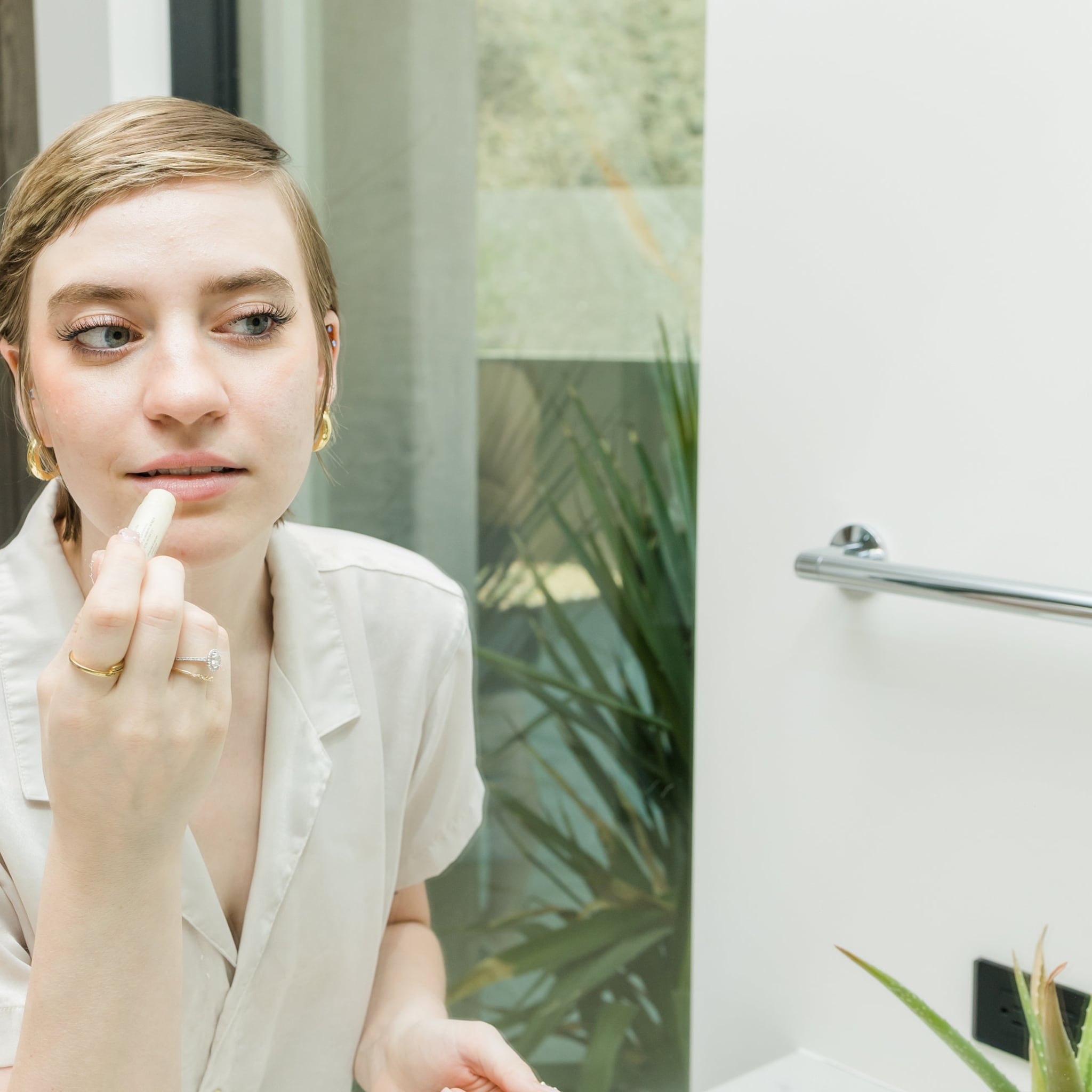 Lip balm application with white tube, fair skin, and gold jewelry in bathroom setting