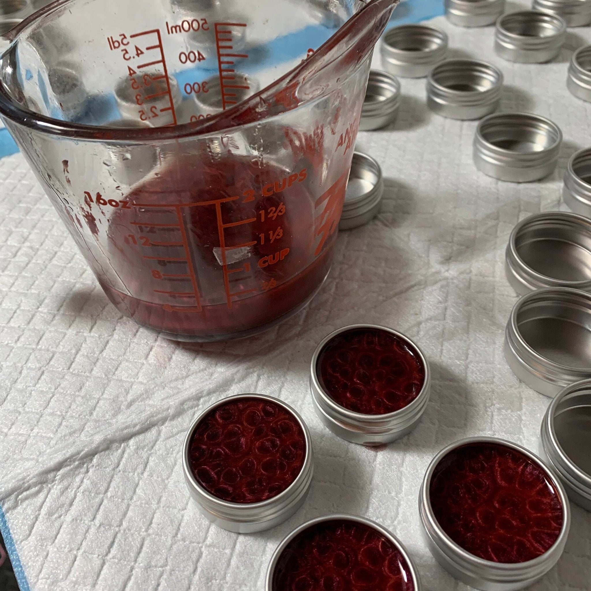 Lip balm filling process with red liquid in measuring cup and small silver containers