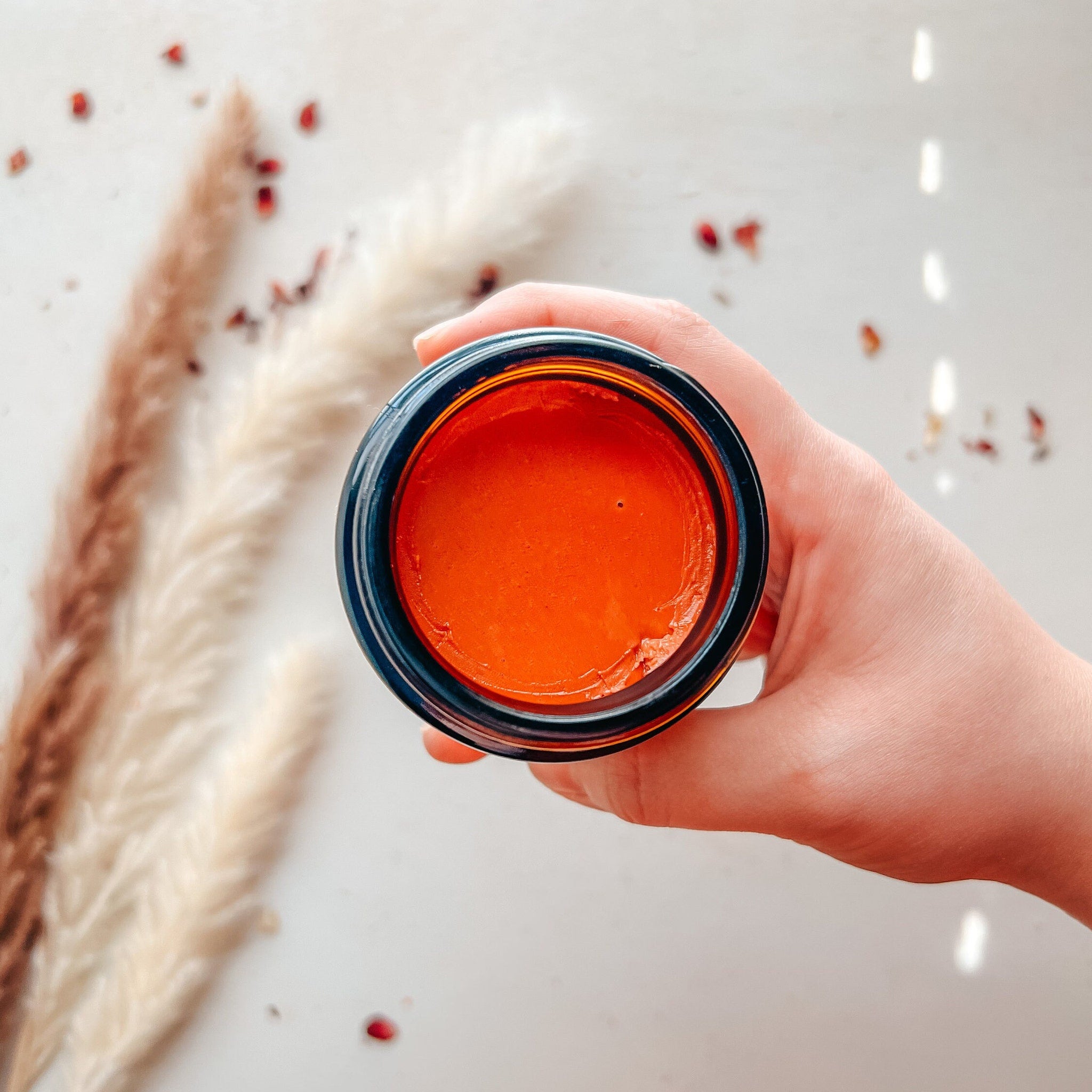 Orange cream in a dark glass jar, held by a hand, natural light and dried flowers