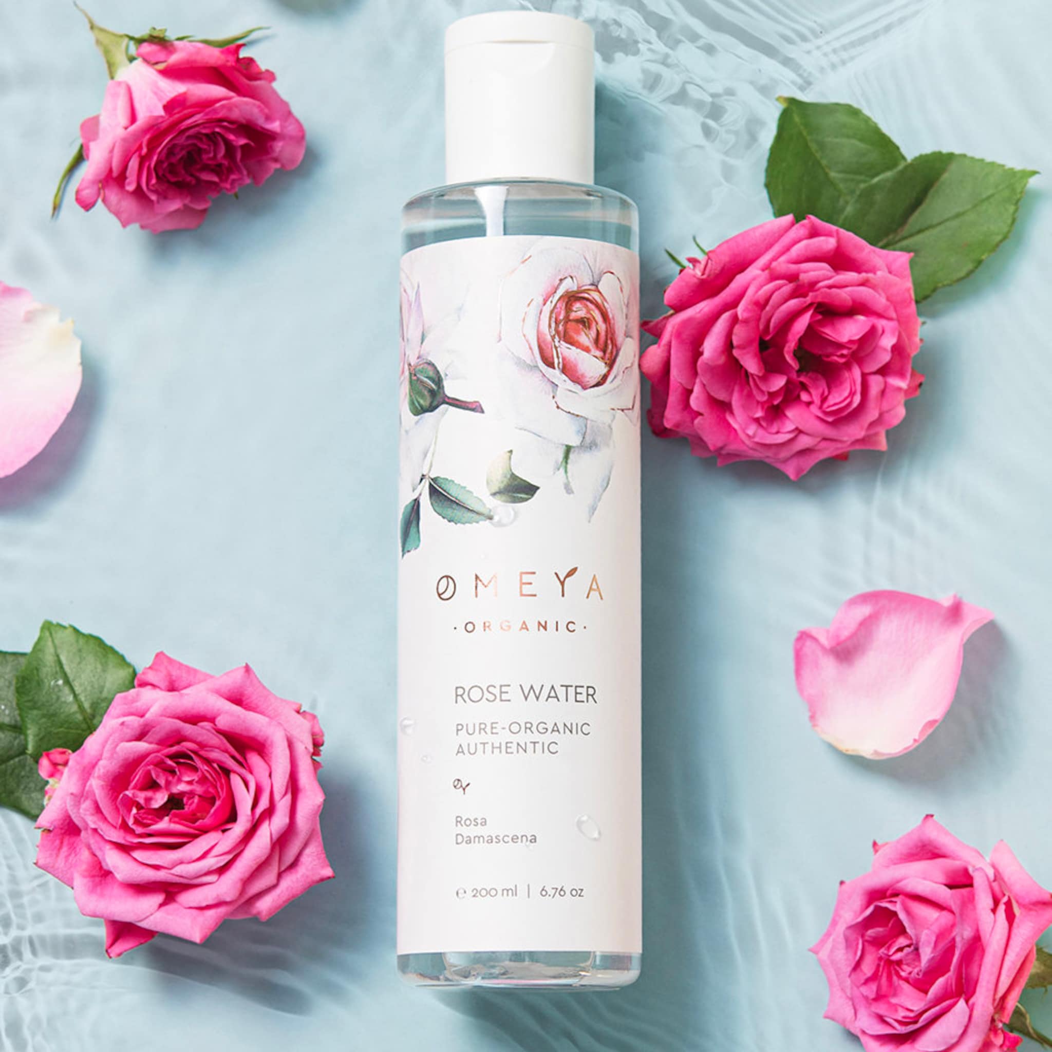 Rose water bottle with pink roses, green leaves, and O MEYA branding on a blue surface