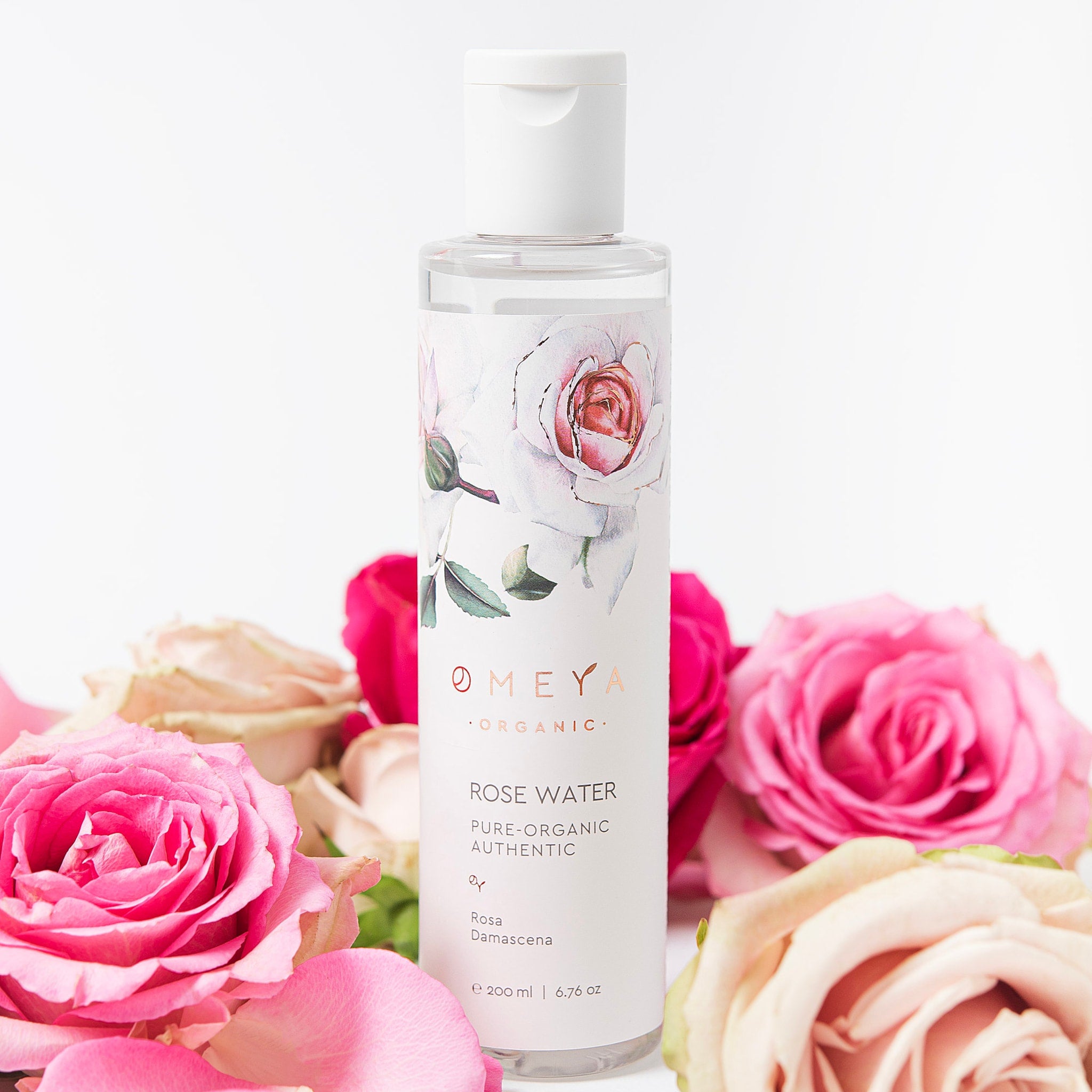 Rose water bottle with white label, floral design, surrounded by pink roses, OMEYA brand visible