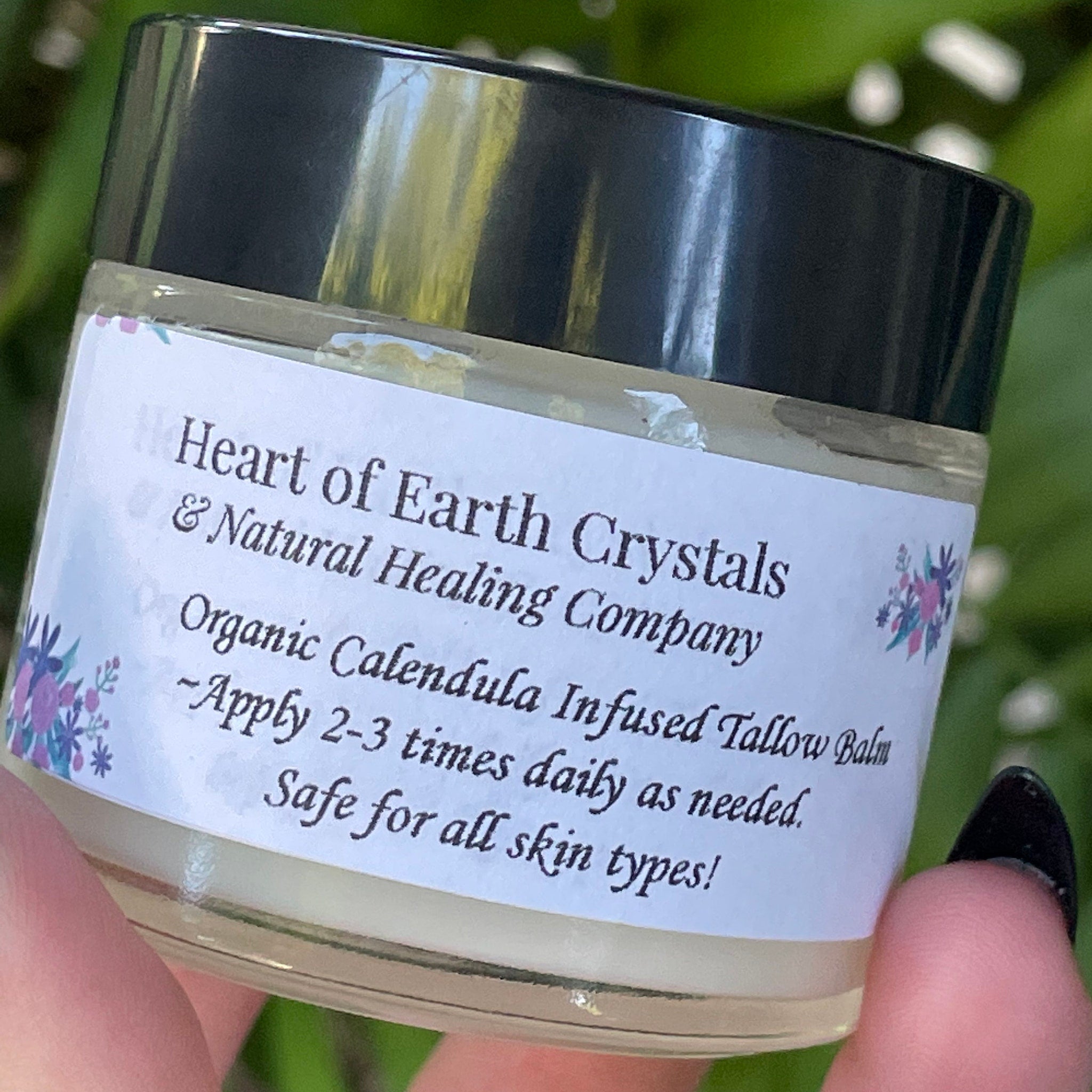 Tallow balm jar with black lid, white label, and floral accents, held in hand