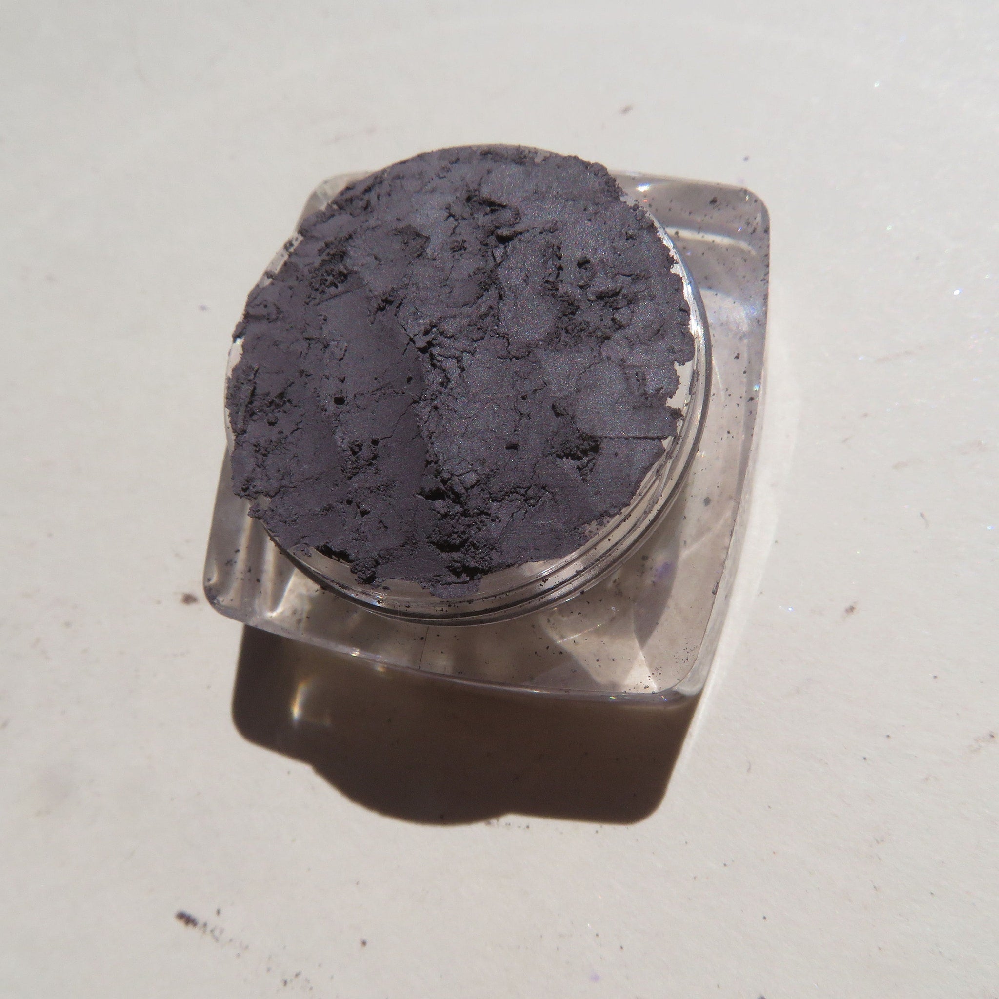 Eyeshadow powder in dark gray color, textured surface, in a clear plastic jar container