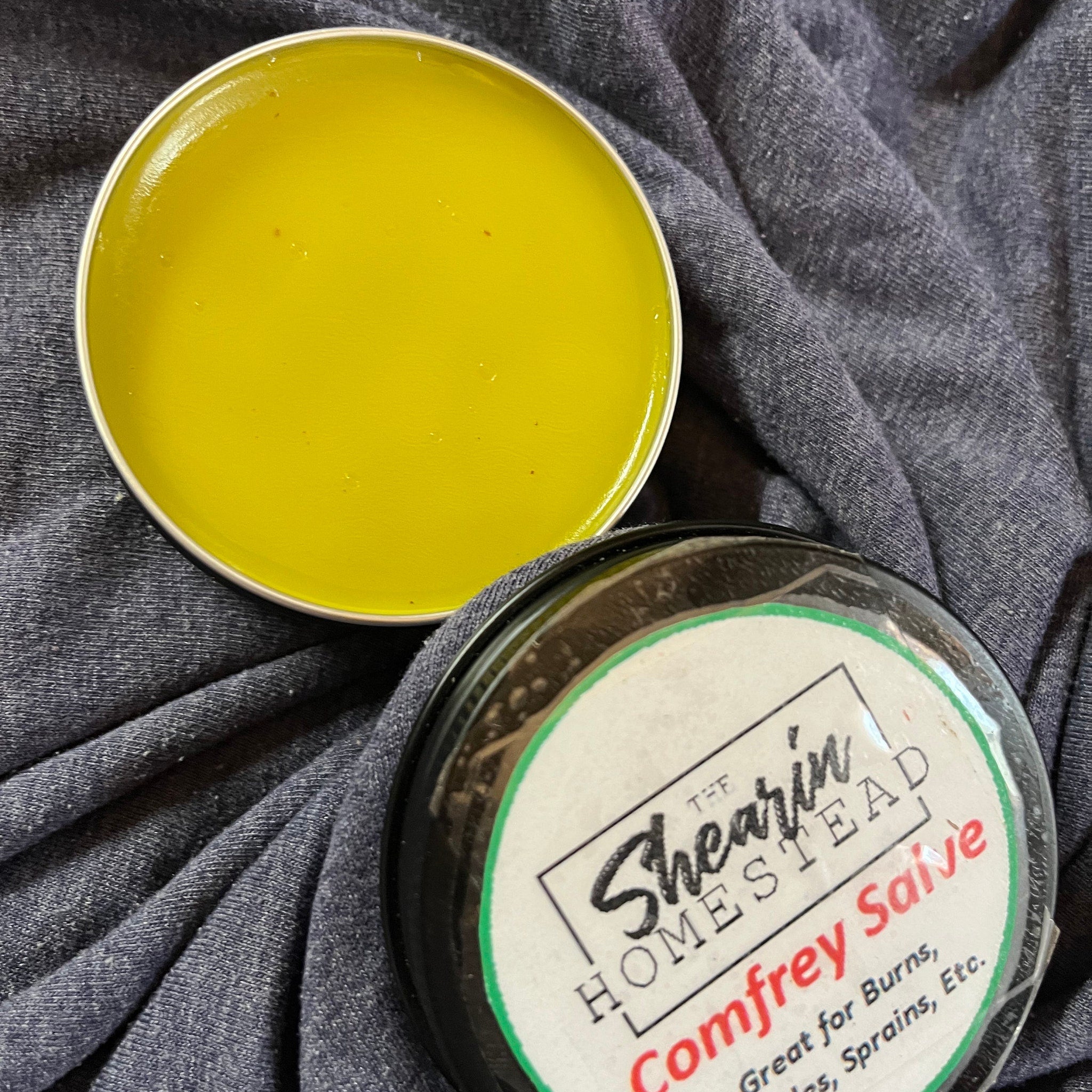 Salve tin with yellow balm, silver rim, and The Shearin Homestead label on gray fabric background