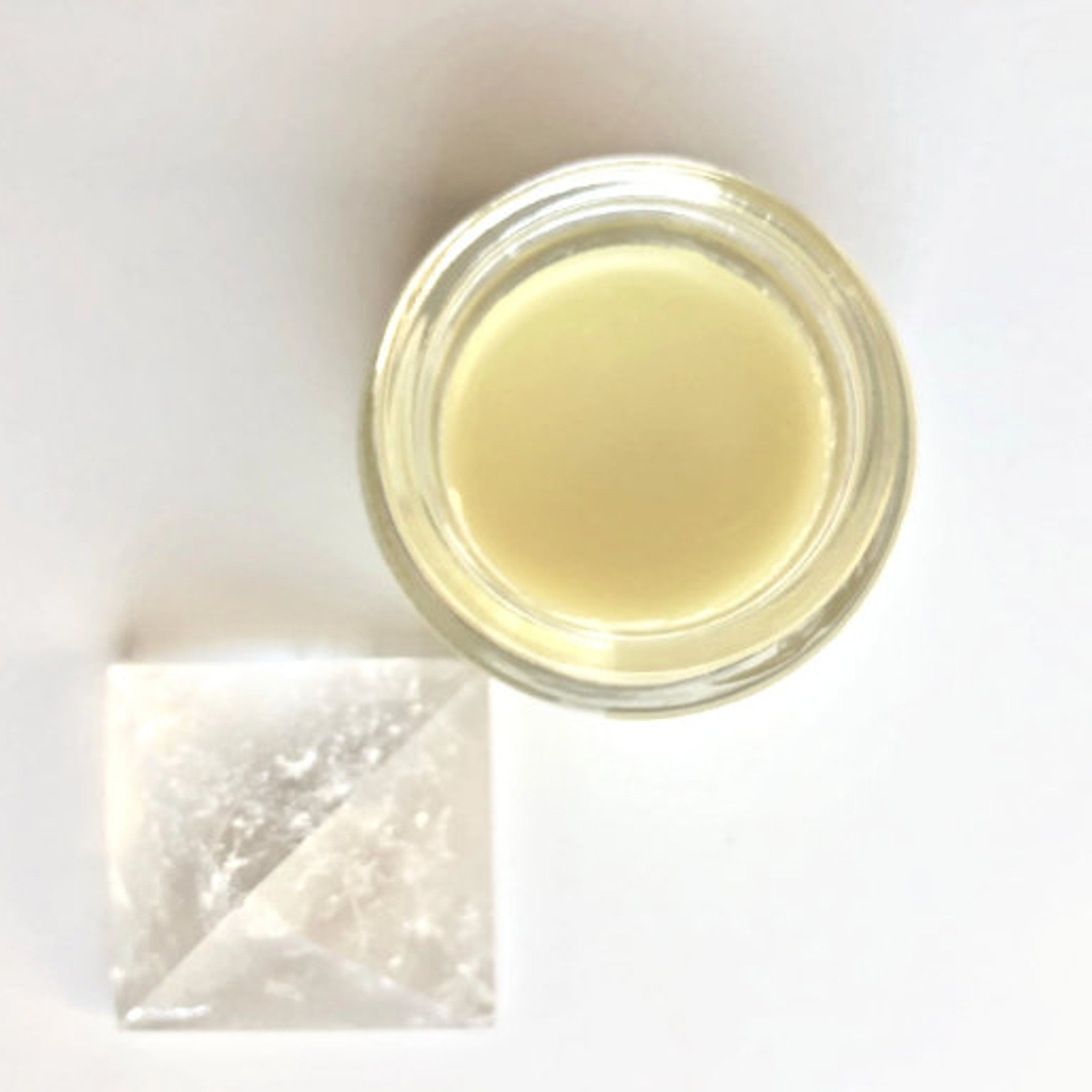 Creamy yellow balm in a clear glass jar, next to a clear crystal pyramid on white surface
