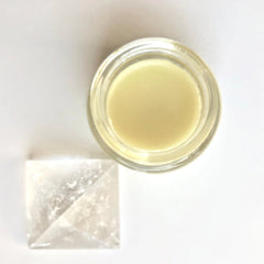 Creamy yellow balm in a clear glass jar, next to a clear crystal pyramid on white surface